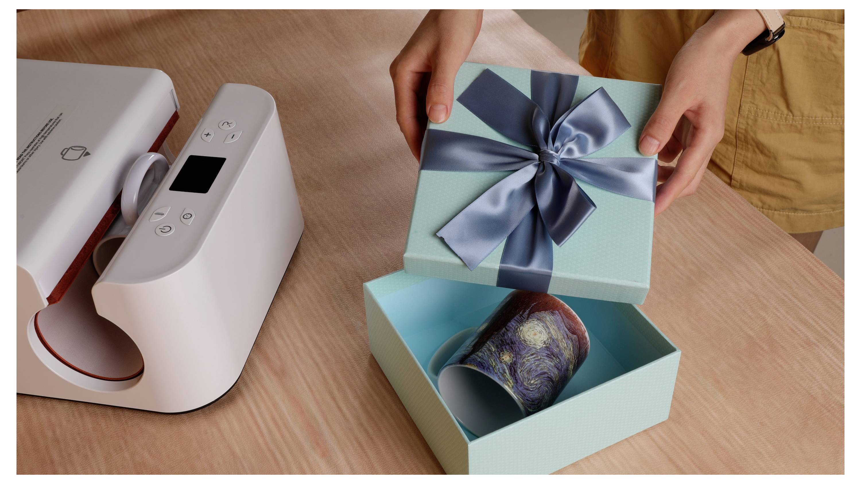 Eine Person öffnet eine blaugrüne Geschenkbox mit einem blauen Band und enthüllt eine Tasse im Inneren, die auf einem Tisch neben einer Wärmepresse platziert ist.