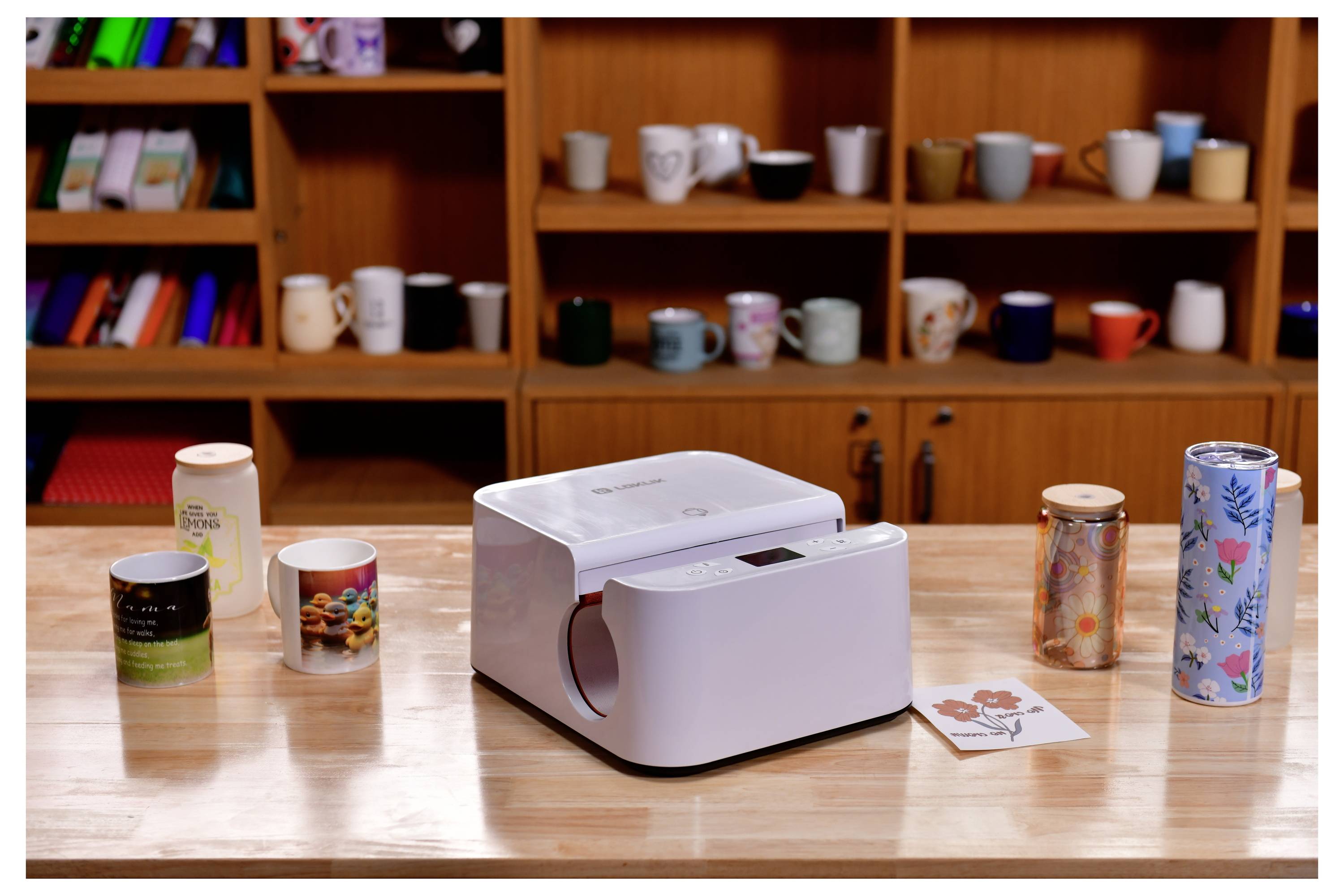 Ein Mugdruckmaschine auf einem Tisch, umgeben von verschiedenen Bechern und Tassen, mit Regalen voller zusätzlicher Becher im Hintergrund.