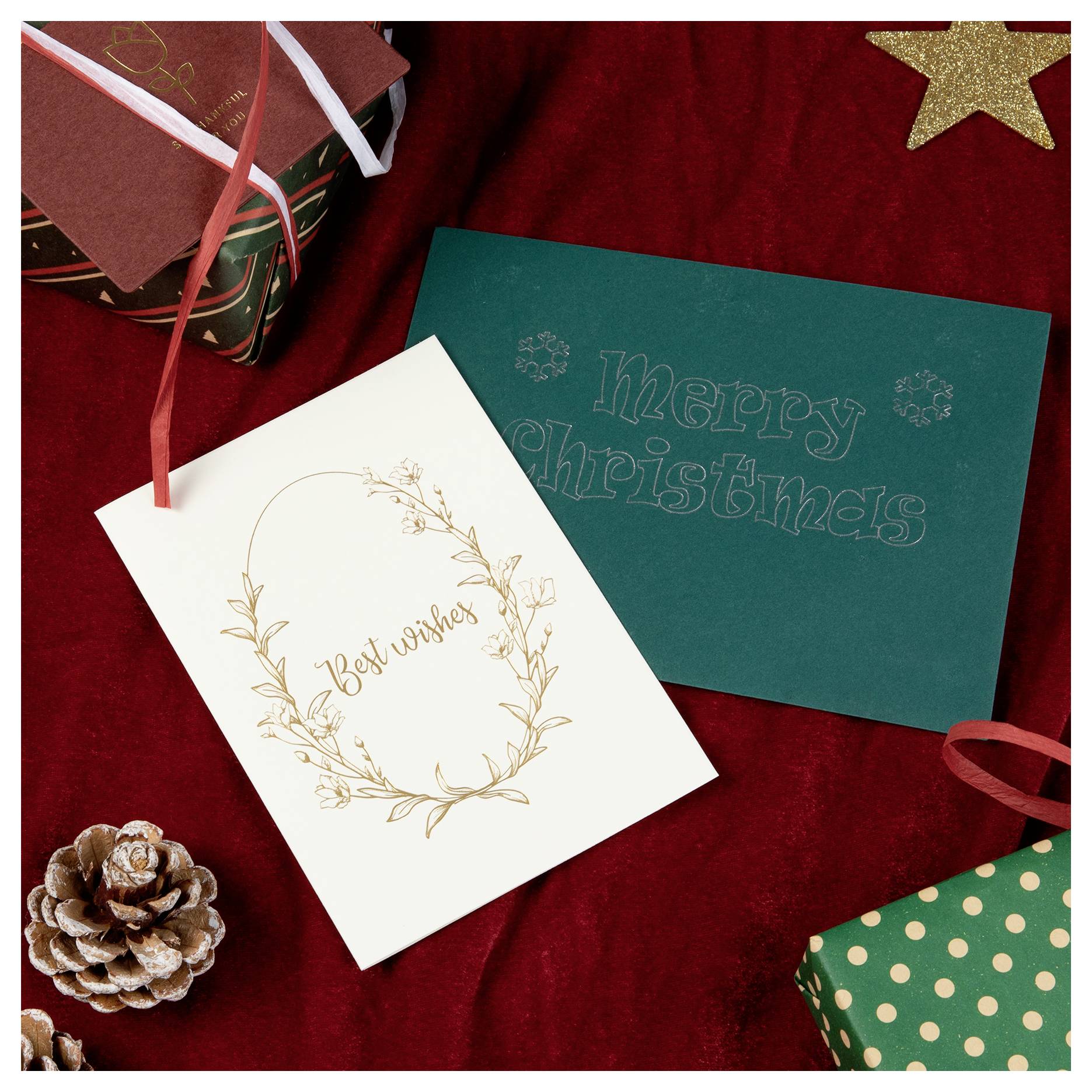 Weihnachtskarte umgeben von Geschenken und einer Tannenzapfen auf rotem Samtuntergrund. Die grüne Karte zeigt die Aufschrift „Frohe Weihnachten