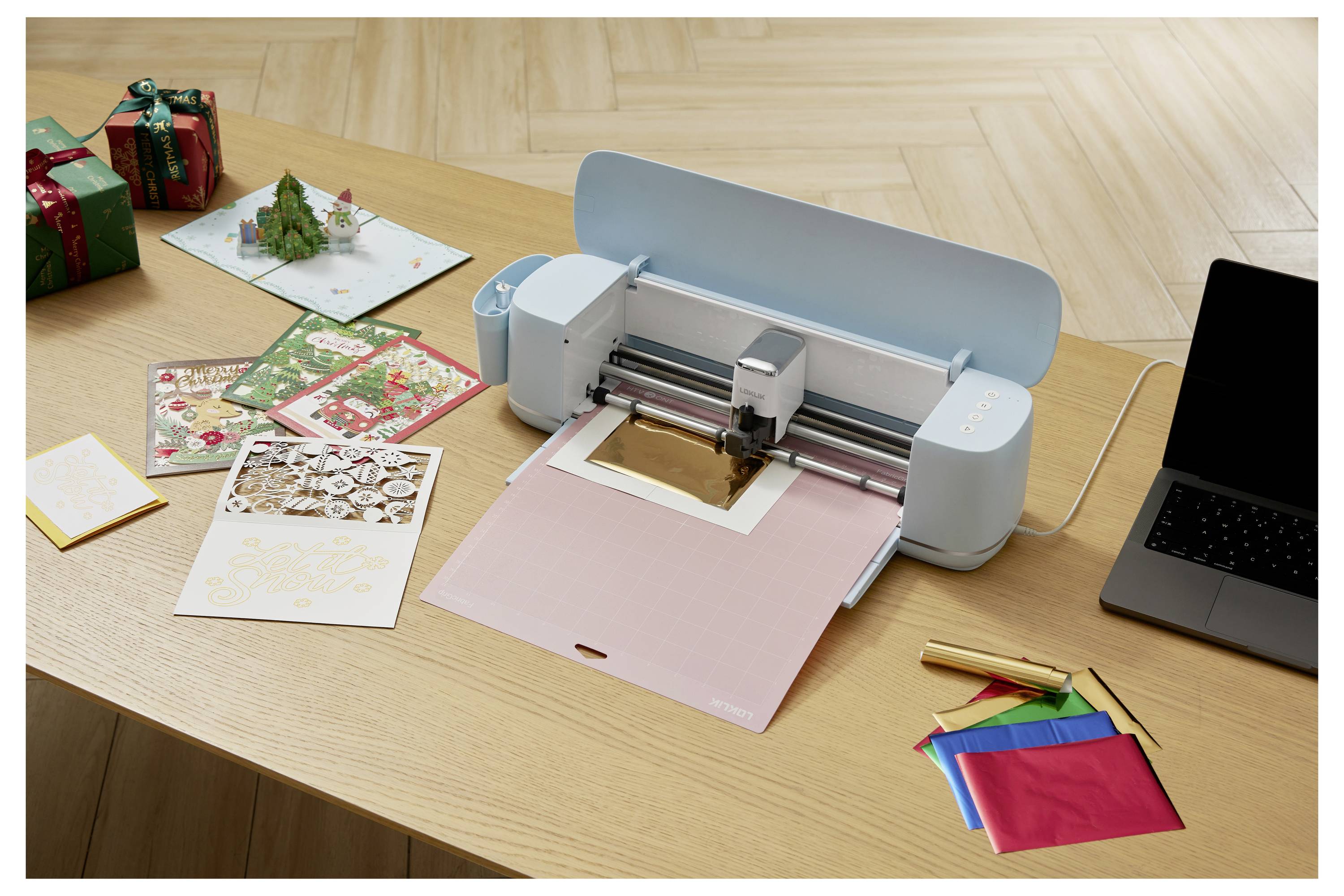 Eine Bastelmaschine schneidet Goldpapier auf einem Tisch mit bunten Materialien, verpackten Geschenken und weihnachtsthematischen Karten in der Nähe.