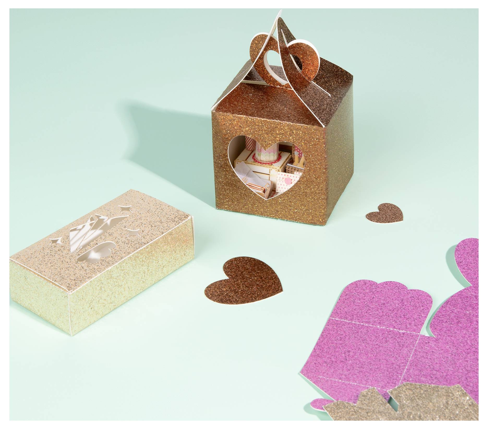 Eine glitzernde braune herzförmige Schachtel und ein rosa Ausschnitt auf einer mintgrünen Oberfläche, neben einer geschlossenen beigen Schachtel mit eleganten Details.