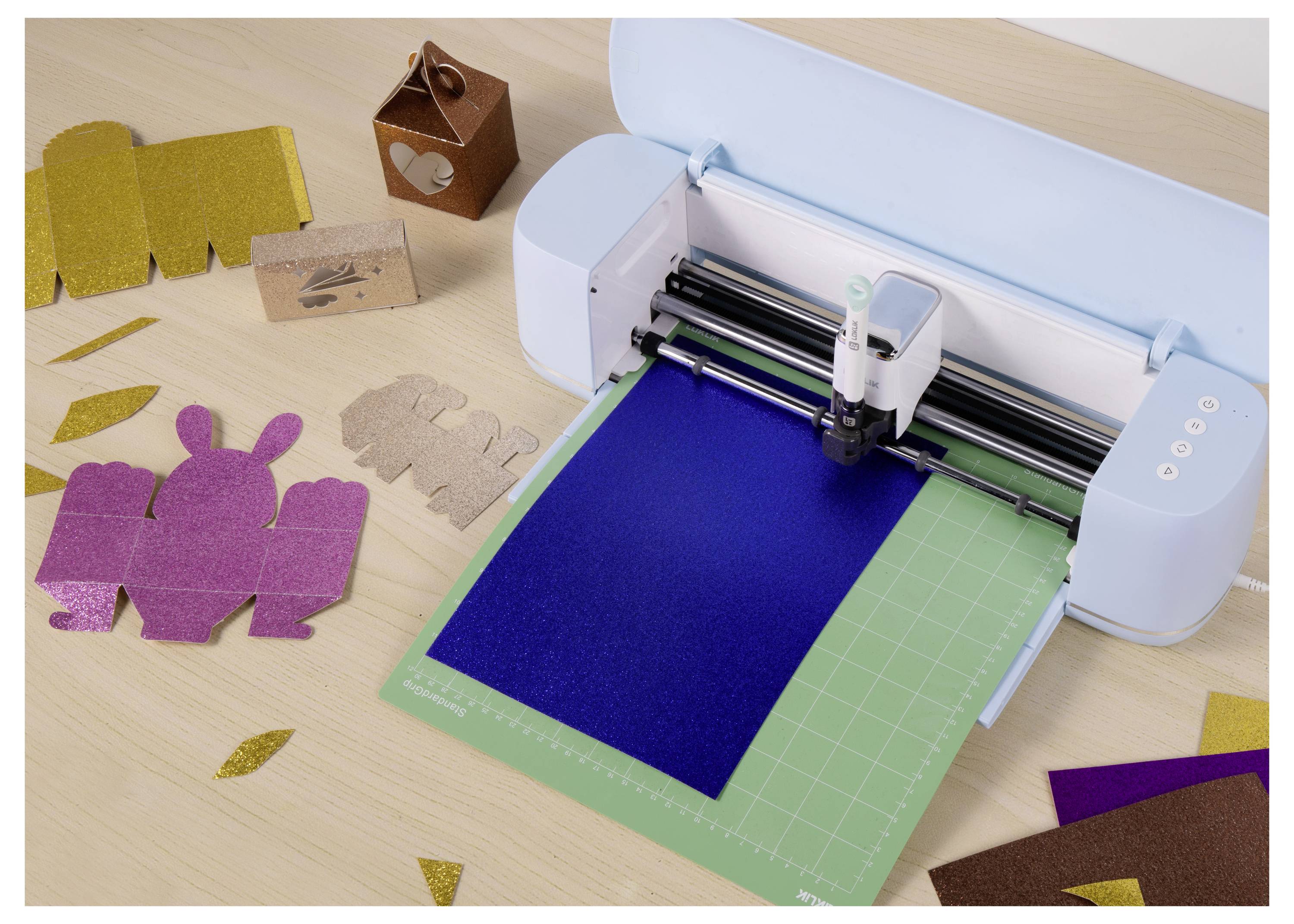 Eine Schneidemaschine in Aktion mit blauem Papier, umgeben von verschiedenen ausgeschnittenen Kartonformen und Designs auf einem Holztisch.