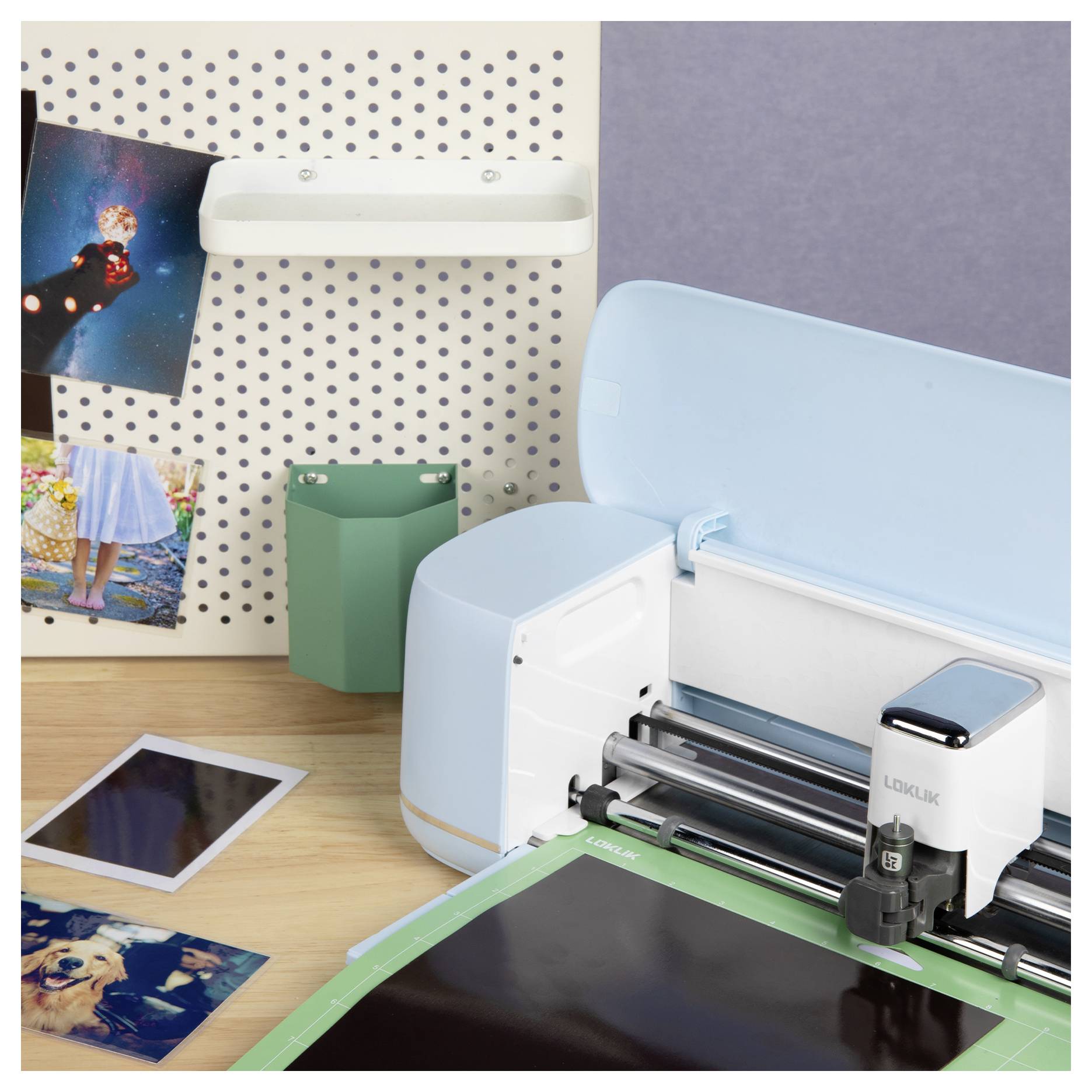 Eine Schneidemaschine auf einem Tisch neben Fotos und Bastelzubehör, mit einer Wandpegplatte, auf der weitere Fotos und ein Stiftehalter angebracht sind.