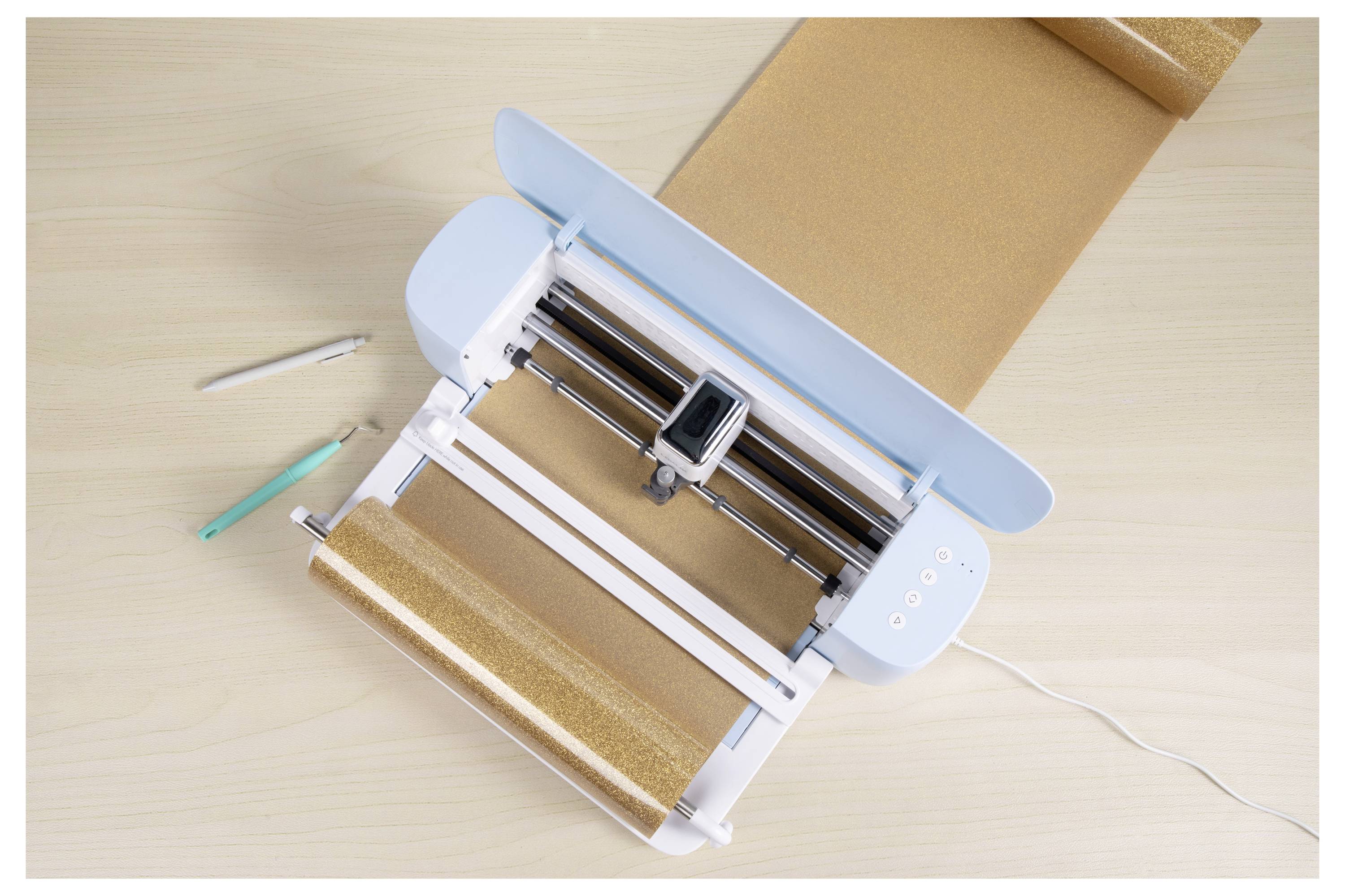 Eine elektronische Schneidemaschine auf einem Holztisch mit einem braunen Material geladen. Neben ihr sind eine Schere und ein Schneidewerkzeug platziert.