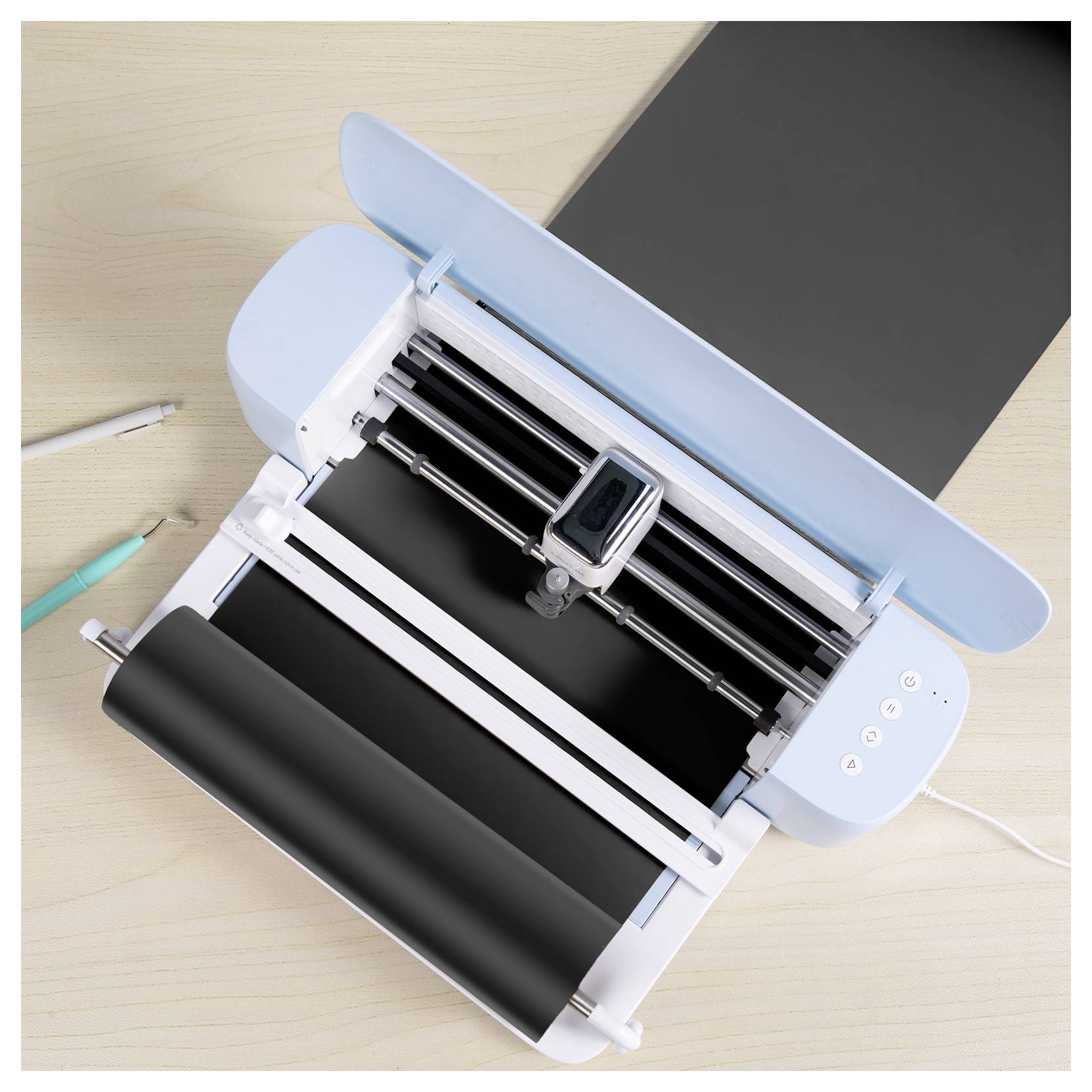 Eine Schneidemaschine mit blauem Deckel schneidet aktiv schwarzes Vinyl auf einem hellem Holztisch, mit einem Stift und einem Marker in der Nähe.