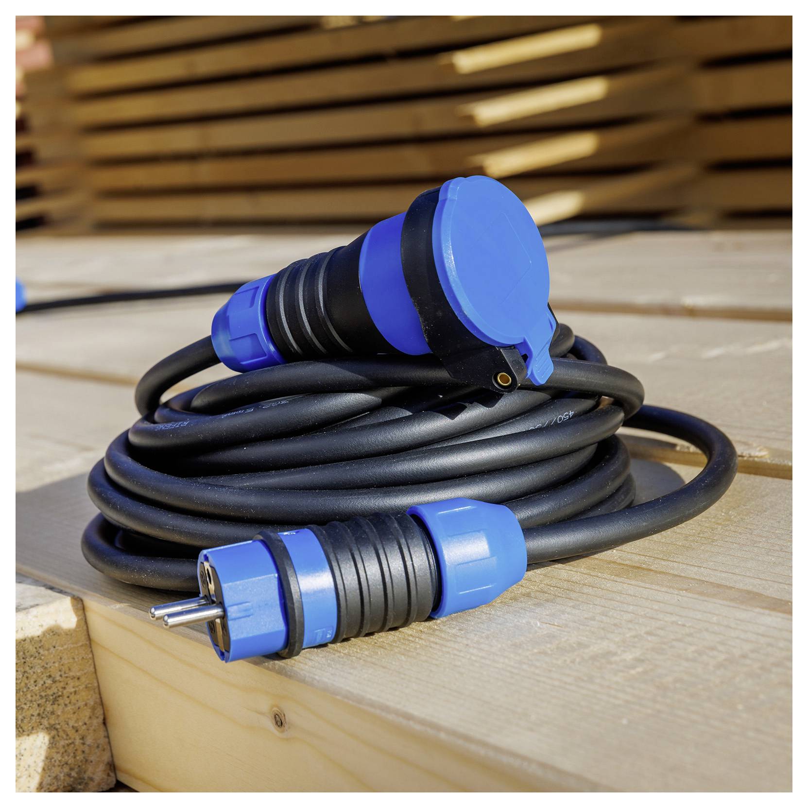 Eine Nahaufnahme eines aufgerollten blauschwarzen Außenverlängerungskabels mit Stecker und Buchse, das auf einer Holzterrasse liegt. Es wird von Sonnenlicht beleuchtet.