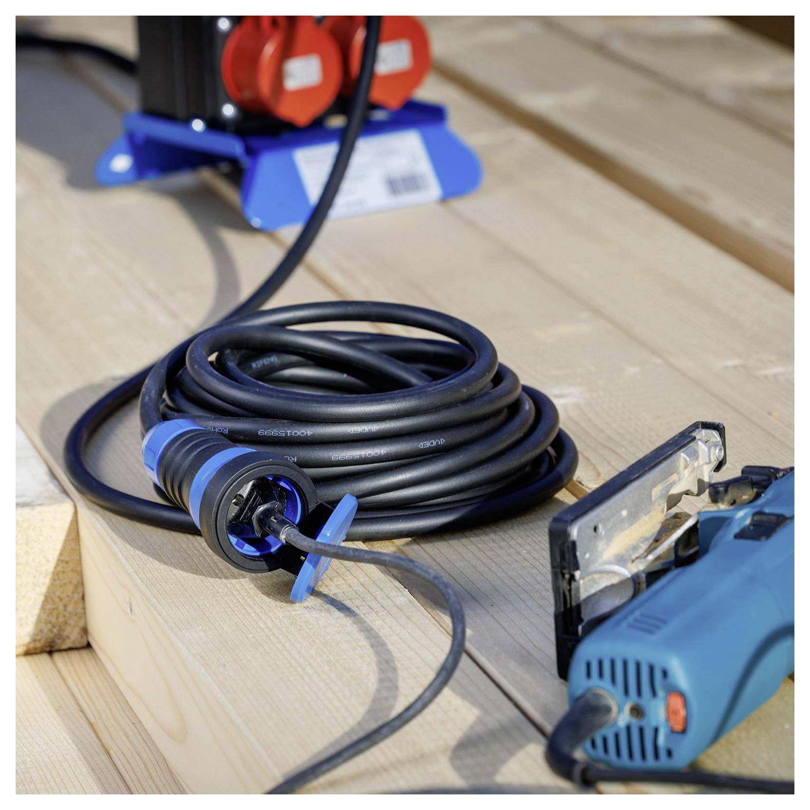 Ein aufgerolltes schwarzes Stromkabel auf einer Holzplattform, angeschlossen an eine blau und rot gefärbte Steckdose. Ein Elektrowerkzeug ist in der Nähe sichtbar.