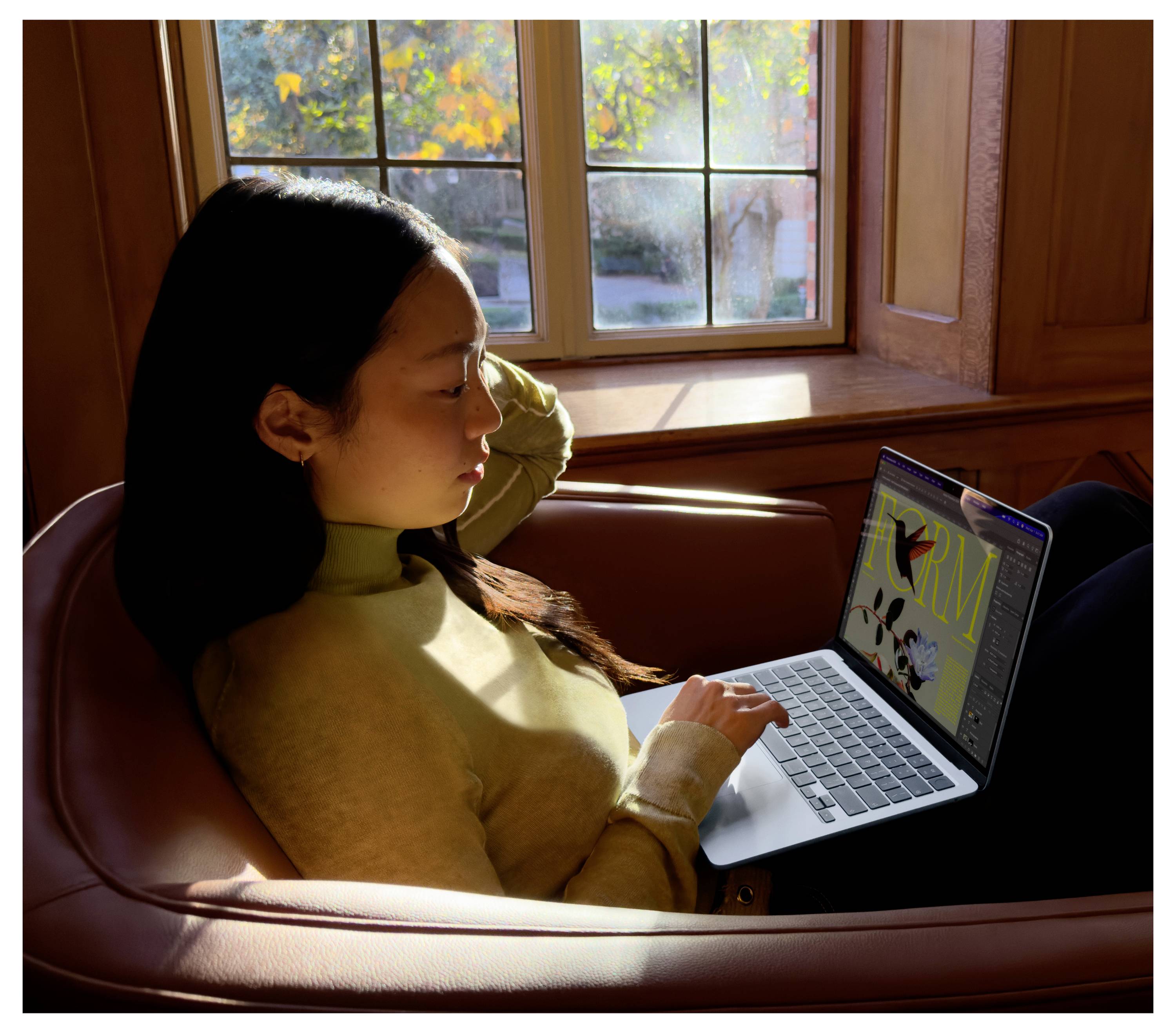 Eine Frau sitzt auf einem Sessel und schaut auf ihren Laptop. Durch das Fenster hinter ihr fällt Sonnenlicht in den Raum.