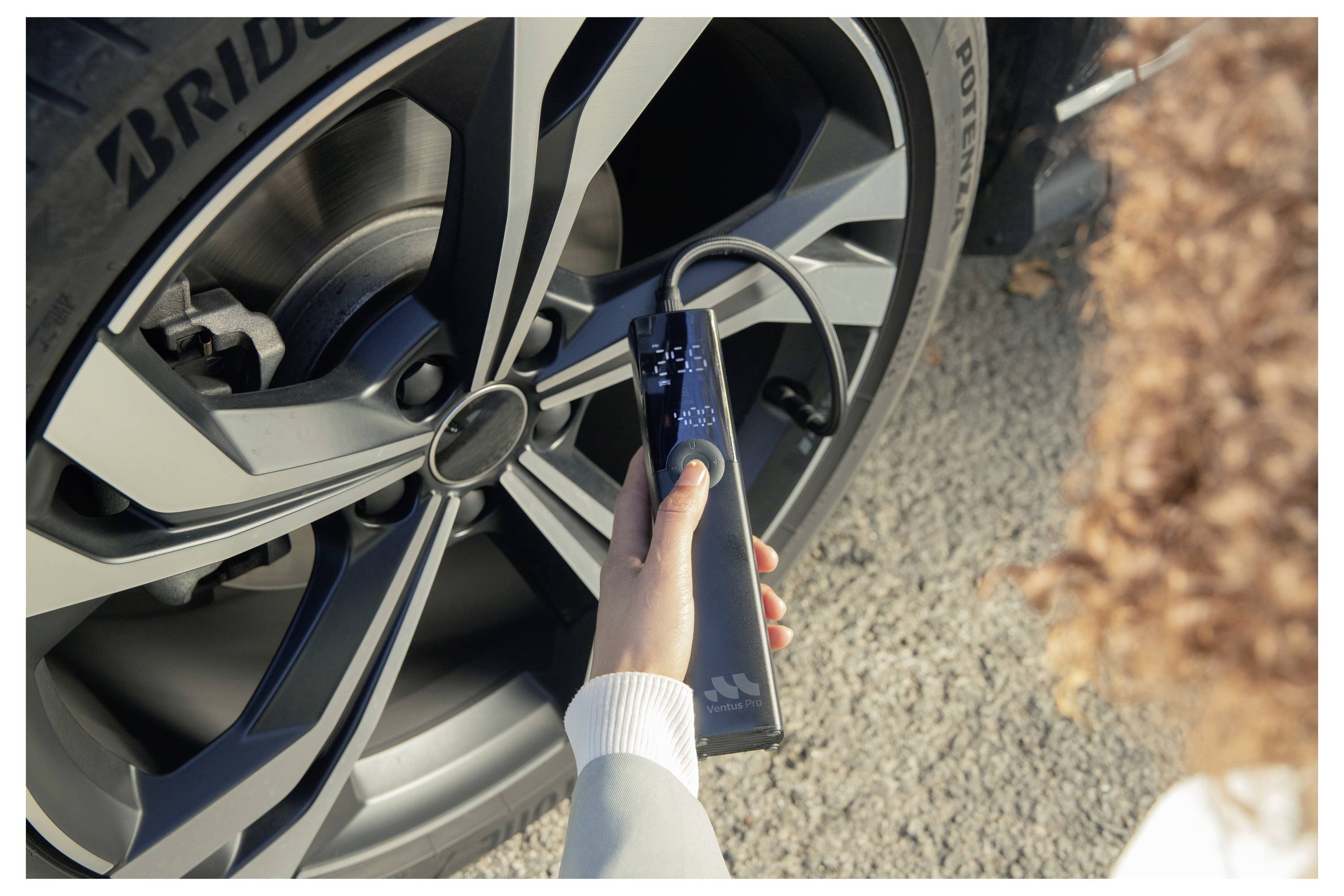 Person, die ein tragbares Gerät in der Nähe eines Autoreifens benutzt, wahrscheinlich zur Inspektion der Reifenprofiltiefe oder Ausrichtung im Freien auf Asphalt.