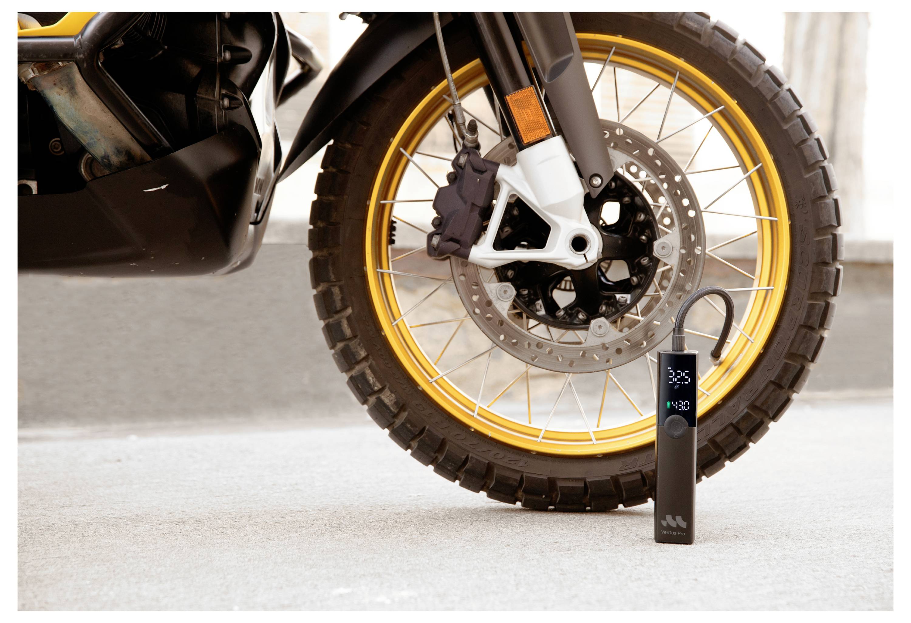 Ein kleines schwarzes digitales Reifendruckmessgerät steht neben dem Vorderrad eines Motorrads mit einer goldenen Felge und einem Stollenreifen.