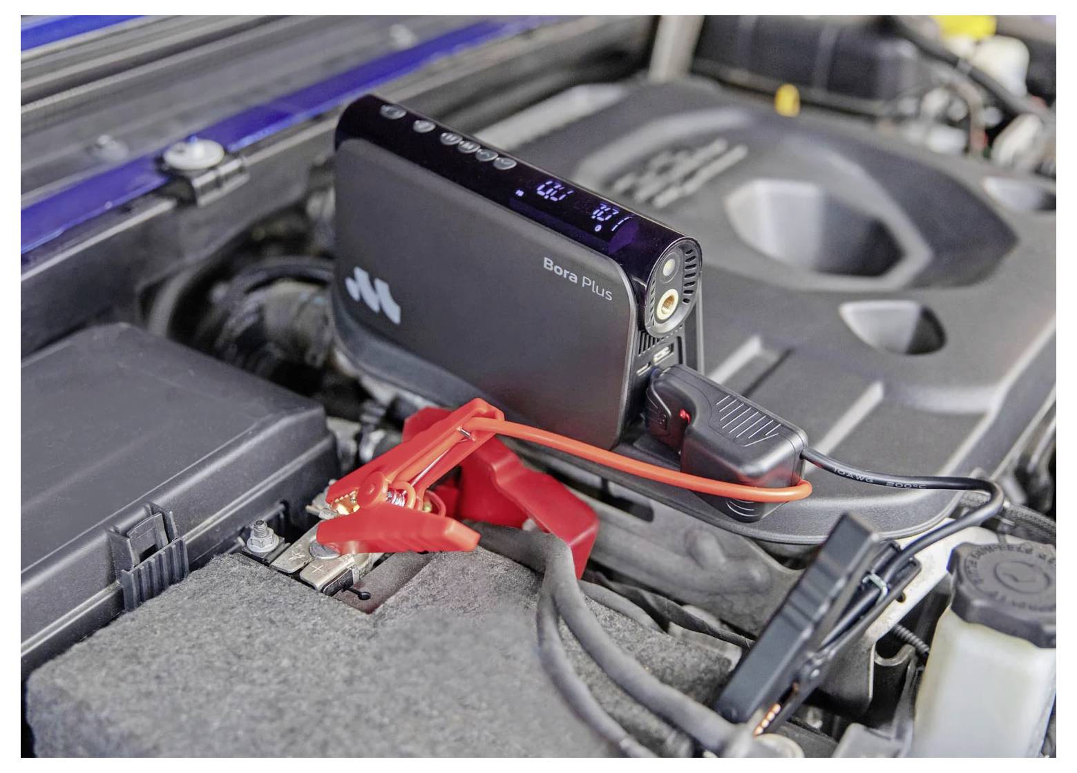 Tragbarer Starthilfegerät, das an einer Autobatterie im Motorraum angebracht ist und durch rote und schwarze Kabel verbunden ist.