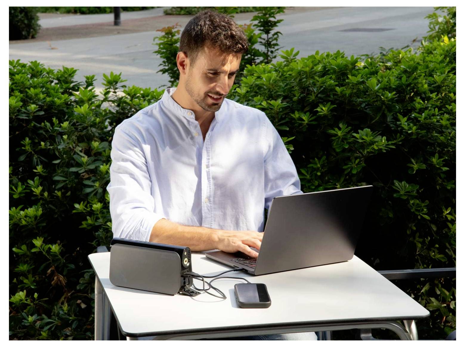 Mann in einem weißen Hemd, der an einem Laptop an einem Außentisch arbeitet, mit einem Tablet und Smartphone neben ihm.