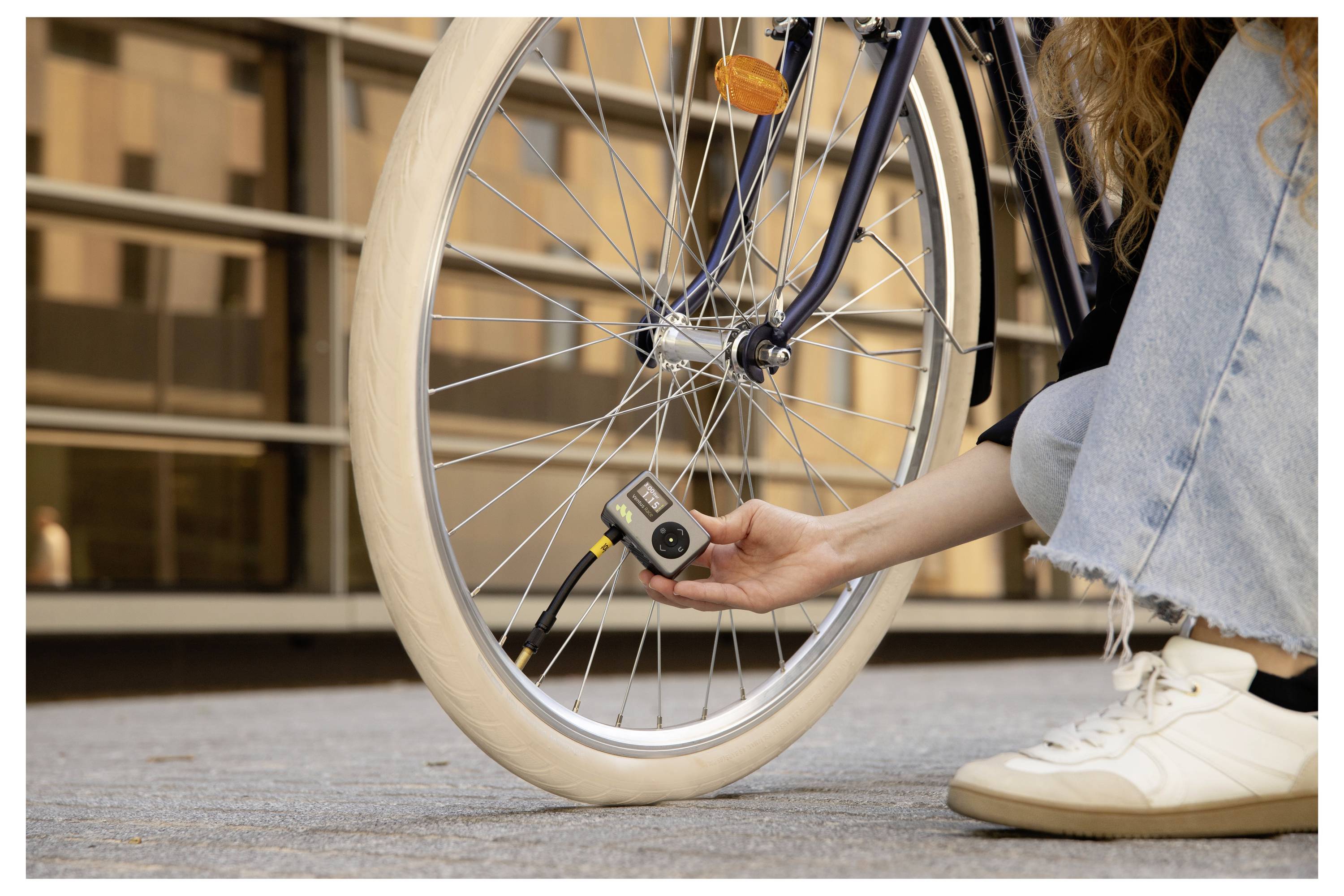 Eine Person in Jeans und Sneakern benutzt ein Druckmessgerät, um den Reifen eines Fahrrads auf einer gepflasterten Fläche vor einem Gebäude zu überprüfen.