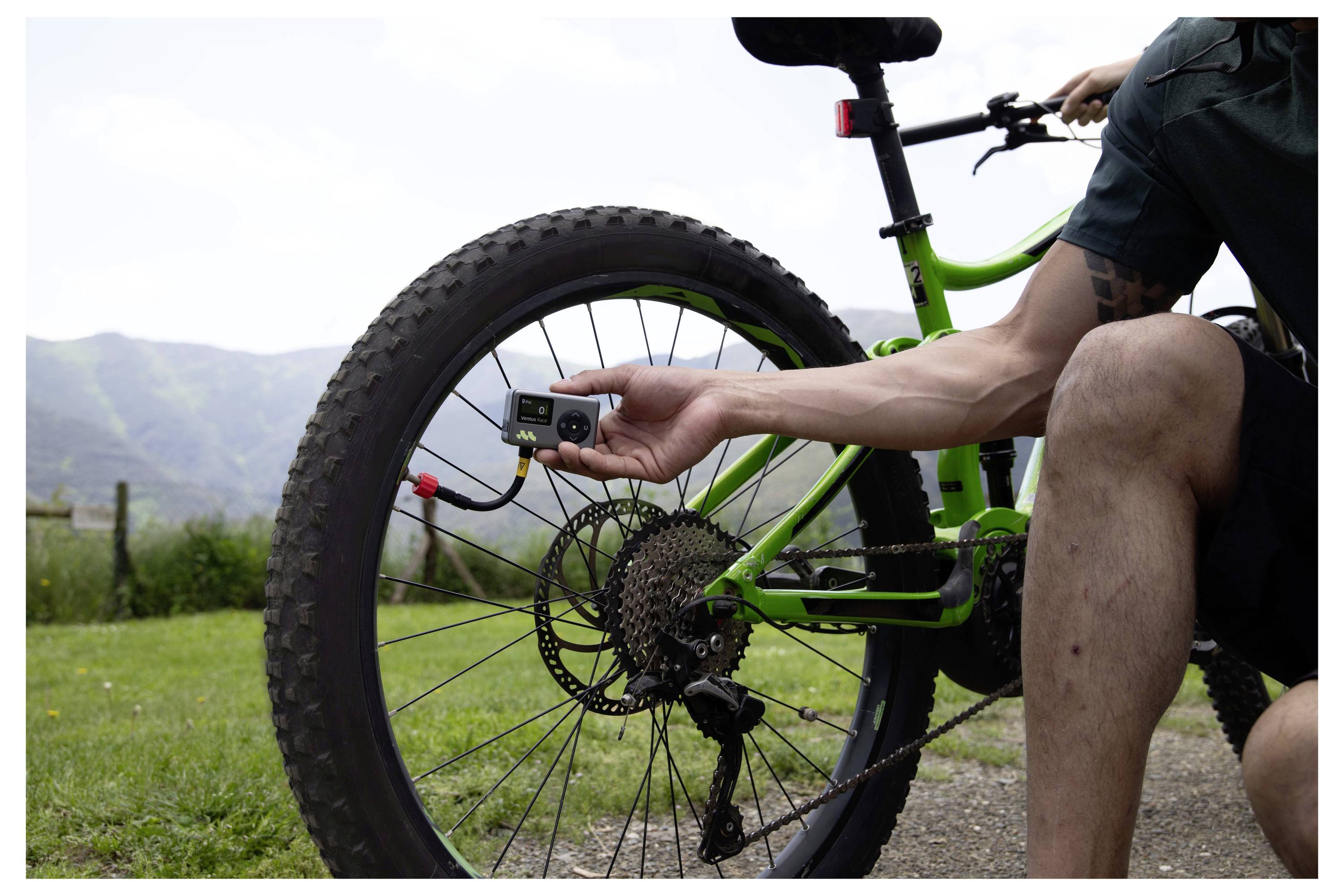 Eine Person auf einem grünen Mountainbike überprüft den Reifendruck des Hinterrads mit einem digitalen Manometer im Freien auf einem grasigen Pfad, mit Hügeln im Hintergrund.