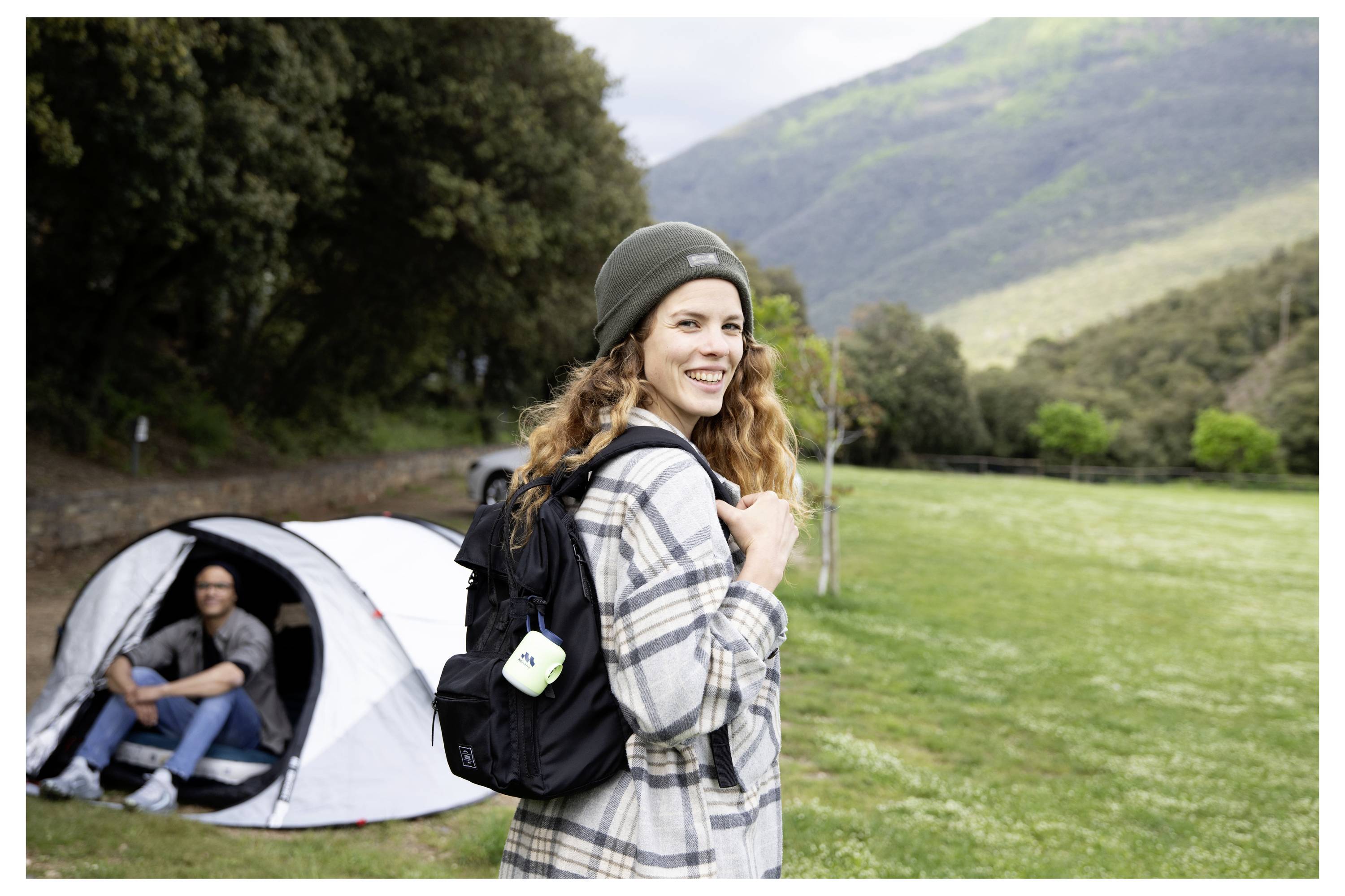 Lächelnde Frau mit Rucksack steht auf einem grasbewachsenen Campingplatz, während ein Mann in einem Zelt sitzt und Berge im Hintergrund zu sehen sind.