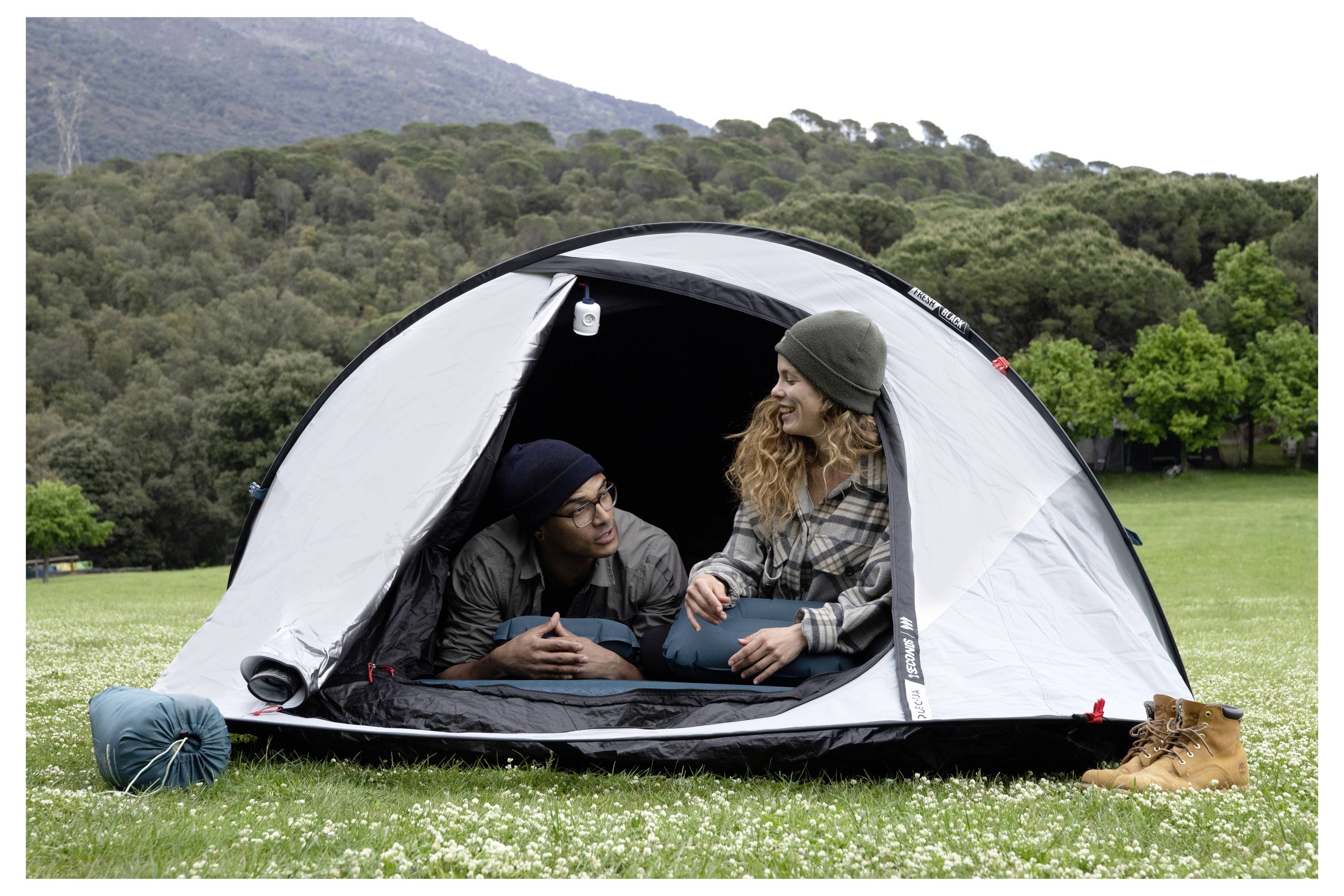 Zwei Camper sitzen in einem grauen Zelt auf einem Wiesenstück, mit einem Schlafsack und Stiefeln draußen, Bergen und Bäumen im Hintergrund.