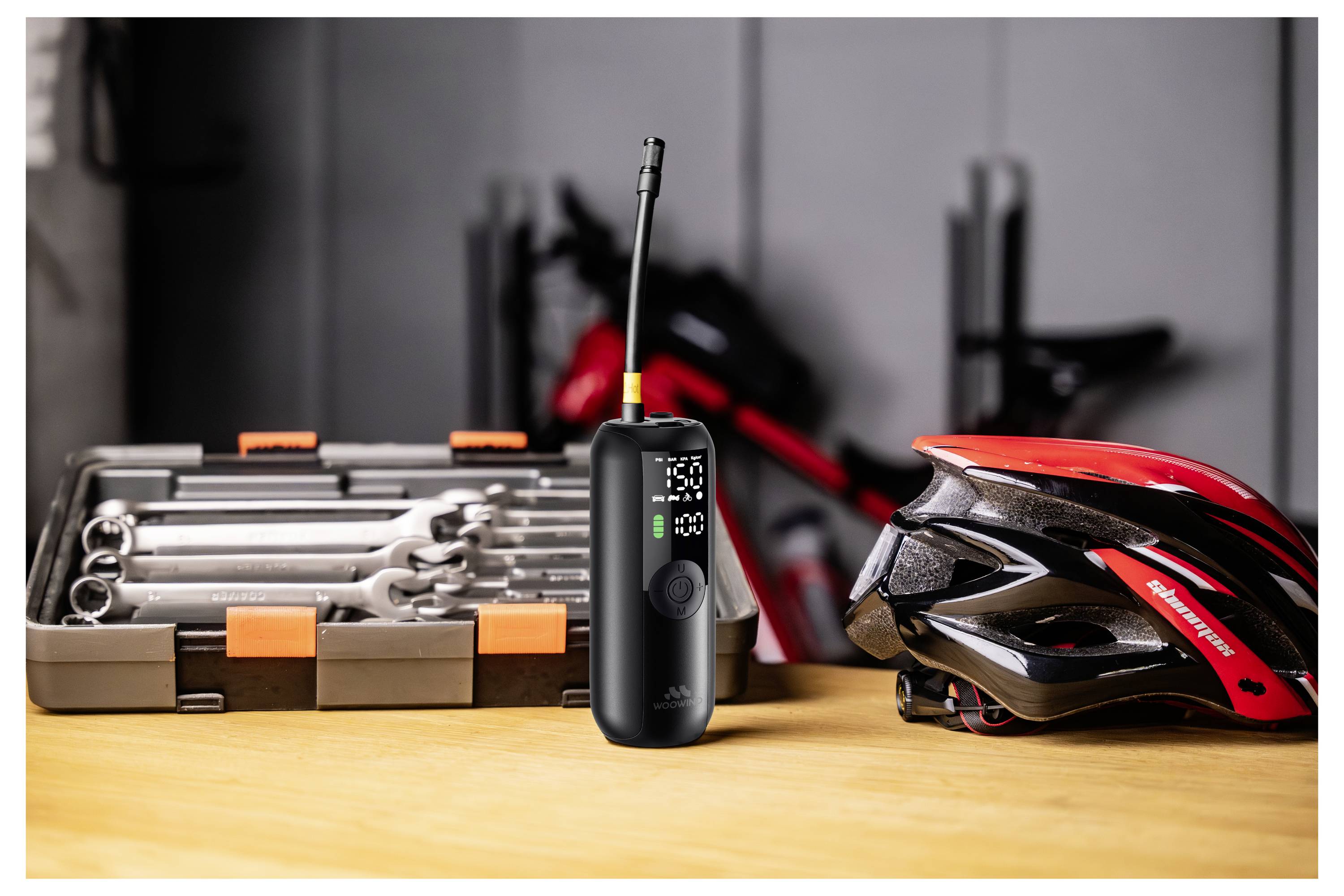 Ein kabelloses Luftpumpen auf einem Tisch neben einem roten Fahrradhelm und einer Werkzeugkiste mit Schraubenschlüsseln, was auf Fahrradwerkzeuge hindeutet.