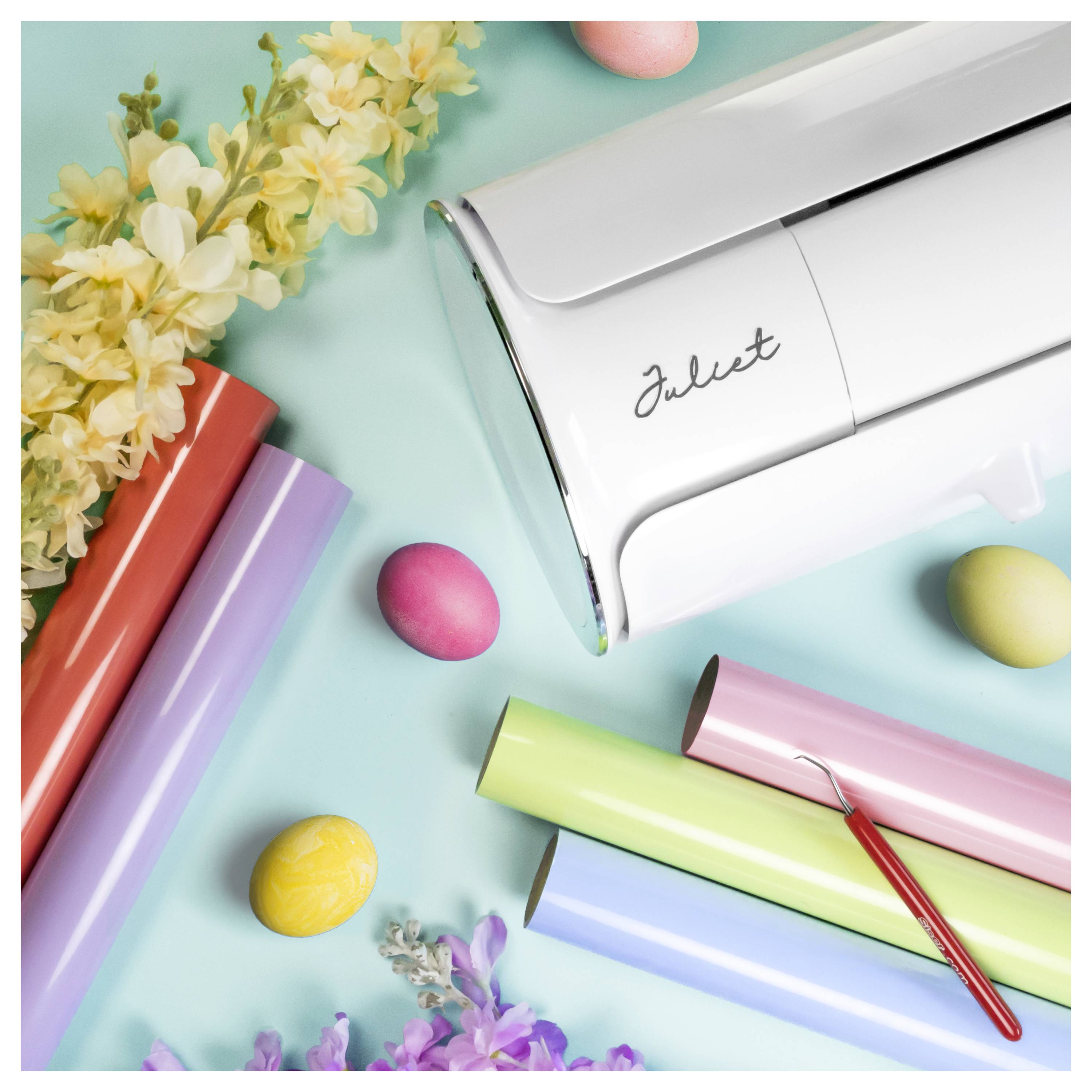 Eine Schneidemaschine umgeben von farbenfrohen Vinyl-Rollen, Blumen, Eiern und einem Hakenwerkzeug, was auf Handwerk und Designaktivitäten hindeutet.