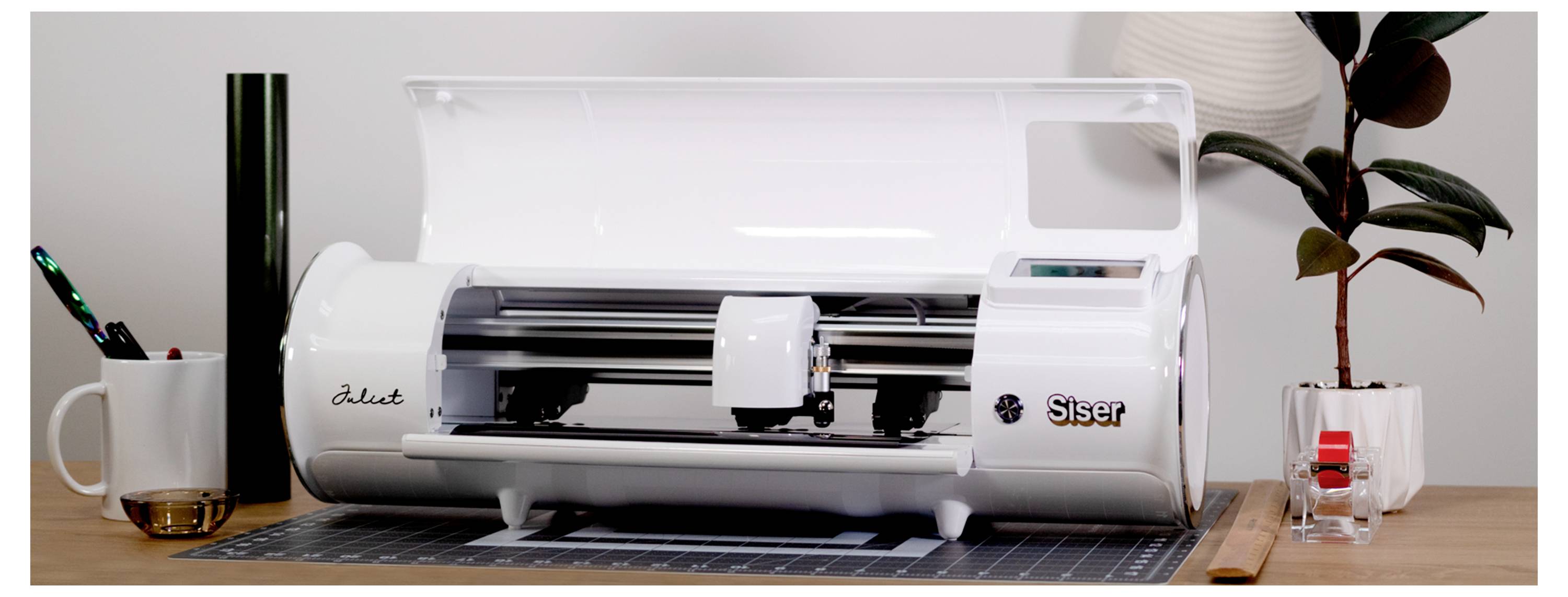 Eine weiße Siser Juliet Schneidemaschine auf einem Holzschreibtisch mit einer Tasse, Stiften, einer schwarzen Vinyl-Rolle und einer kleinen Pflanze in der Nähe.