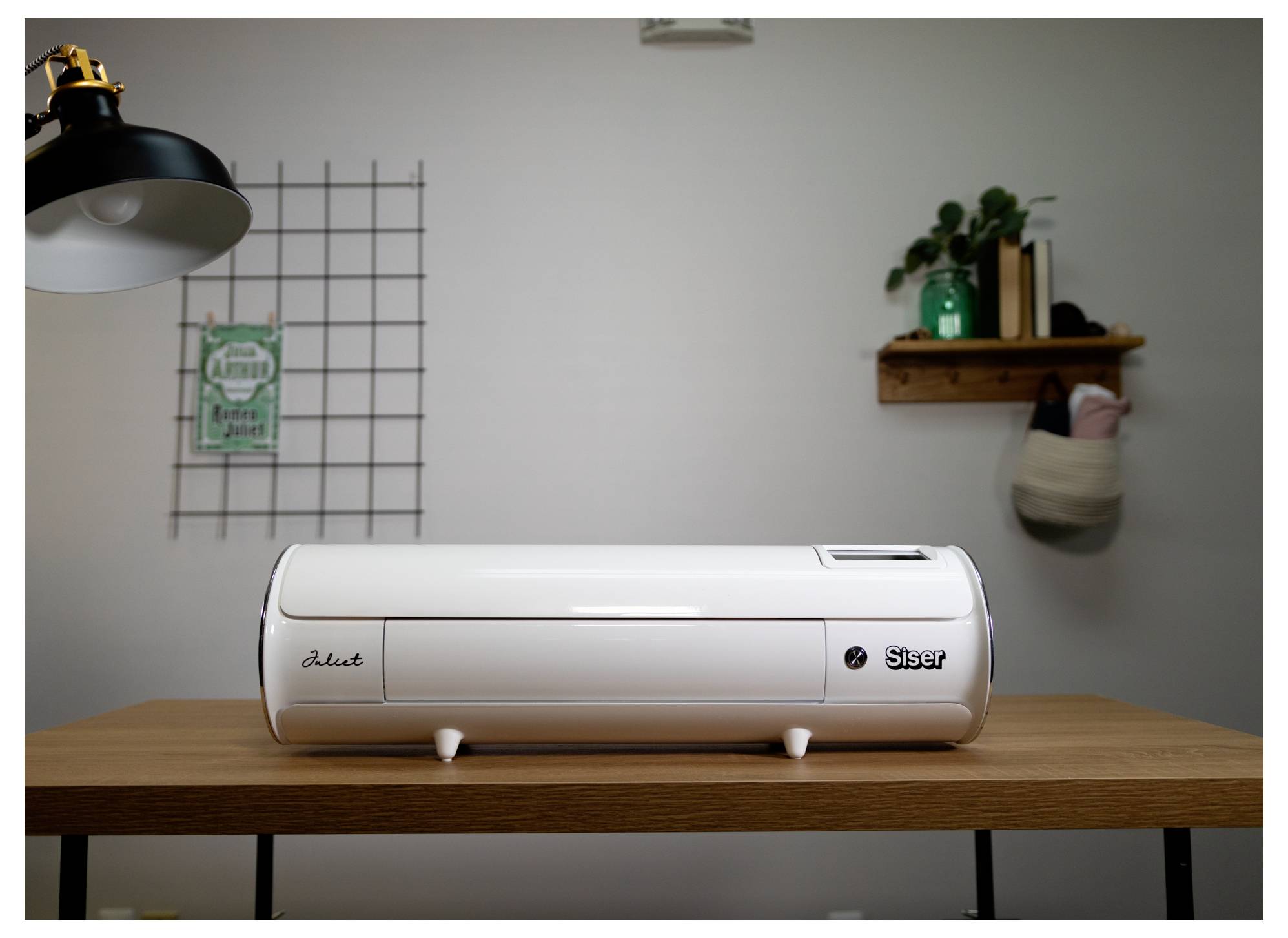 Ein weißer Siser Juliet Schneideplotter sitzt auf einem Holzschreibtisch, mit einer Lampe und Regalen im Hintergrund.