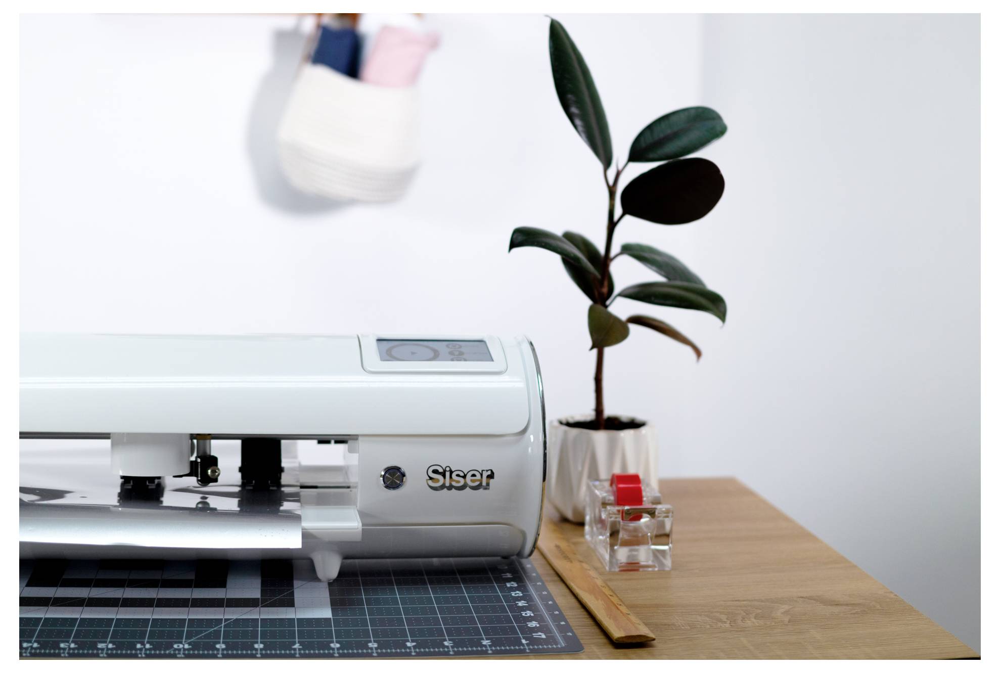 Ein Vinyl-Schneideplotter auf einem Tisch mit einer Pflanze, einer Schneidematte und Messwerkzeugen in der Nähe.