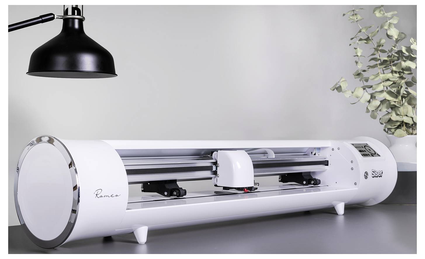 „Eine schlanke, moderne elektronische Schneidemaschine auf einem Tisch, mit einer schwarzen Lampe darüber und Eukalyptuszweigen in der Nähe, in einem hellen Raum.