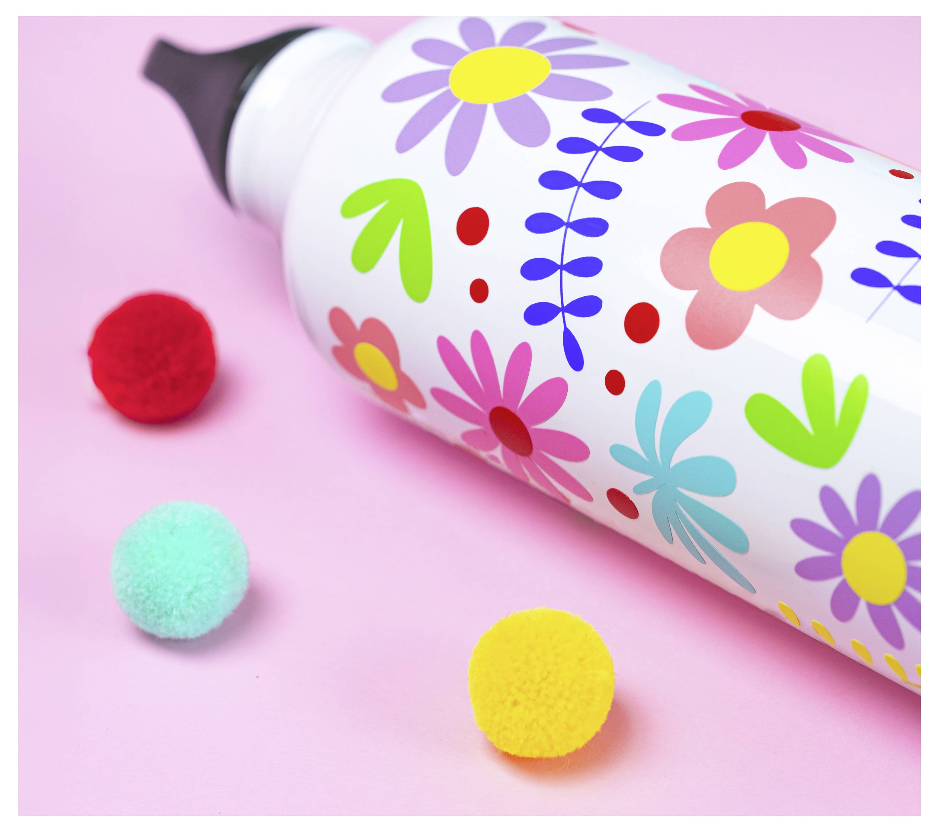 Eine farbenfrohe Wasserflasche mit einem Blumenmuster liegt auf einer rosafarbenen Oberfläche, umgeben von kleinen roten, blauen und gelben Pompons.