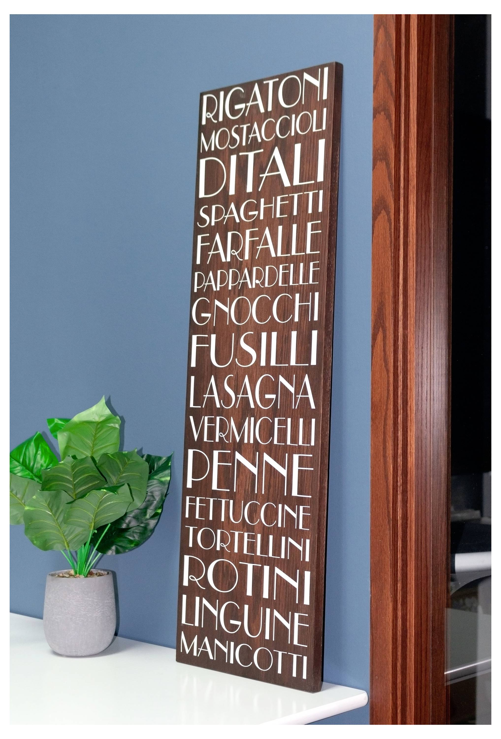 Ein Holzschild listet verschiedene Arten von Nudeln auf, wie 'Rigatoni', 'Spaghetti', 'Fusilli' und 'Linguine', und lehnt an einer blauen Wand neben einer Topfpflanze.