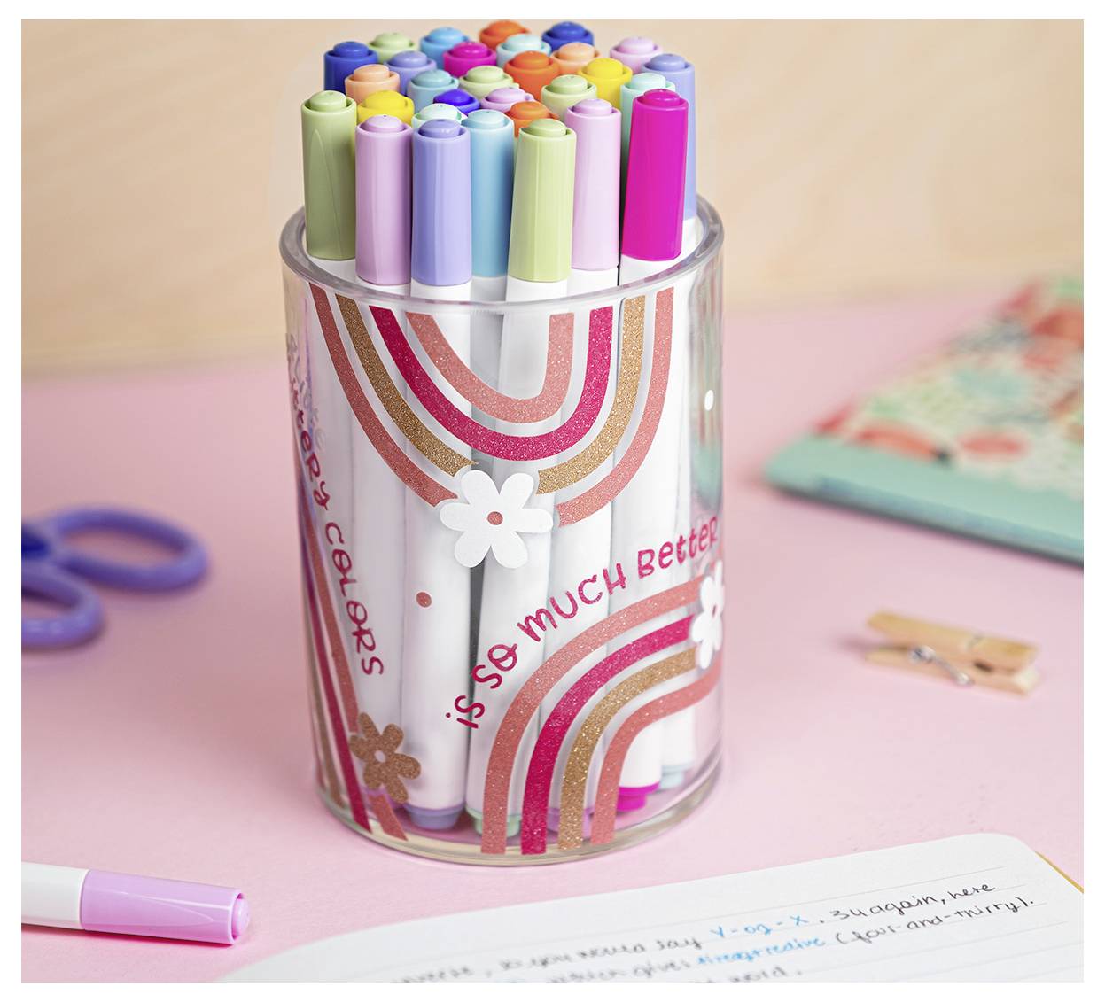 Ein durchsichtiger Becher gefüllt mit verschiedenfarbigen Stiften mit einem Regenbogen- und Blumendesign und dem Text „so much better