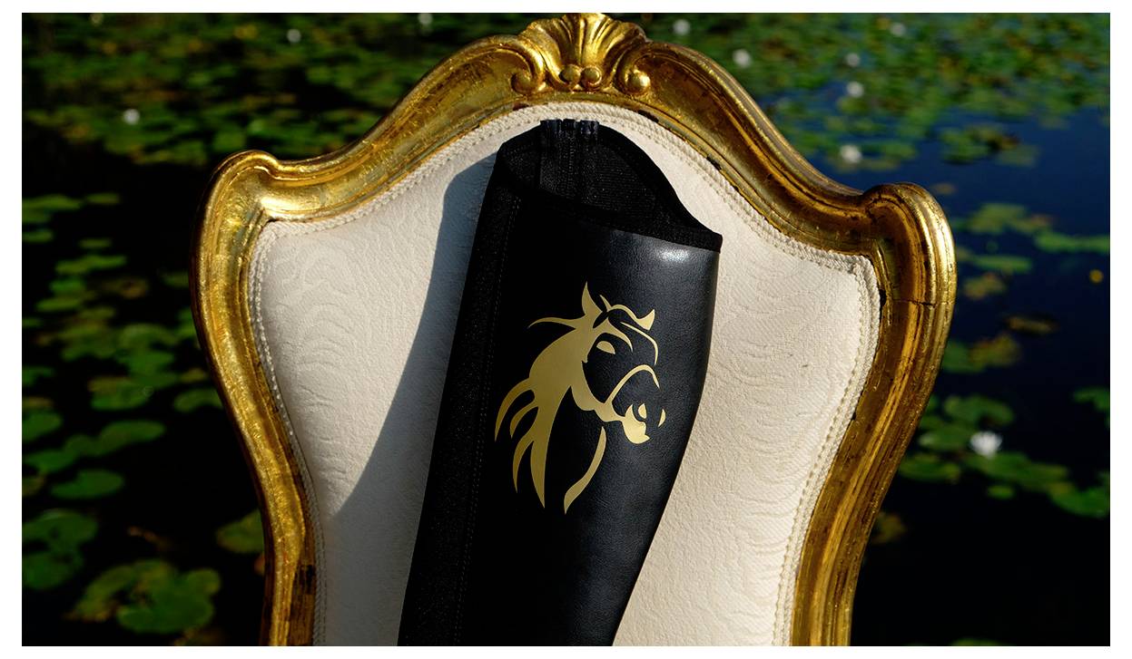 Ein schwarzer Reitstiefel mit einem goldenen Pferdelogo ruht auf einem ornamentalen, weiß-goldenen Stuhl, vor dem Hintergrund eines mit Seerosen bedeckten Teiches.