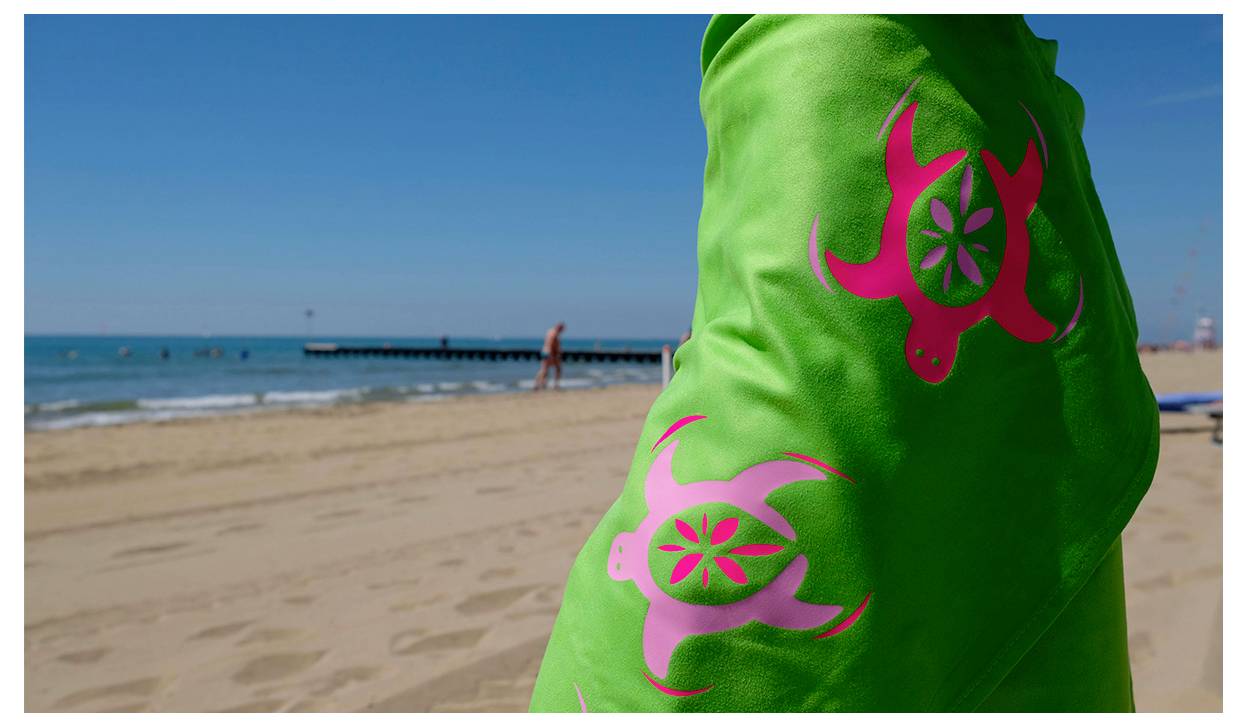 Eine Person, die ein leuchtend grünes Hemd mit rosa Meeresschildkröten-Designs trägt, steht auf einem Sandstrand mit dem Ozean und einem hölzernen Pier im Hintergrund.