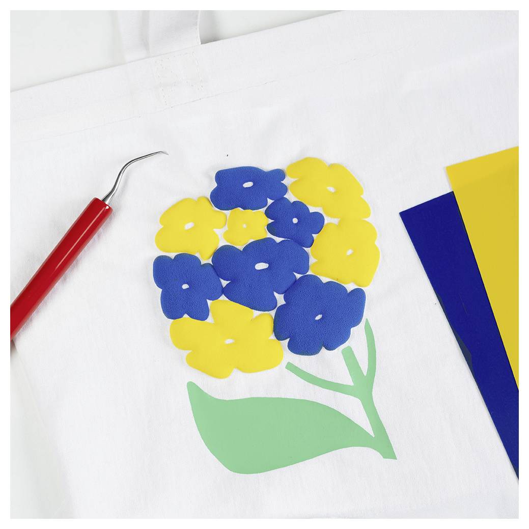 Buntes Blumendesign mit blauen, gelben Blütenblättern und grünen Blättern auf weißem Stoff, neben einem roten Handwerkswerkzeug und farbigem Papier.
