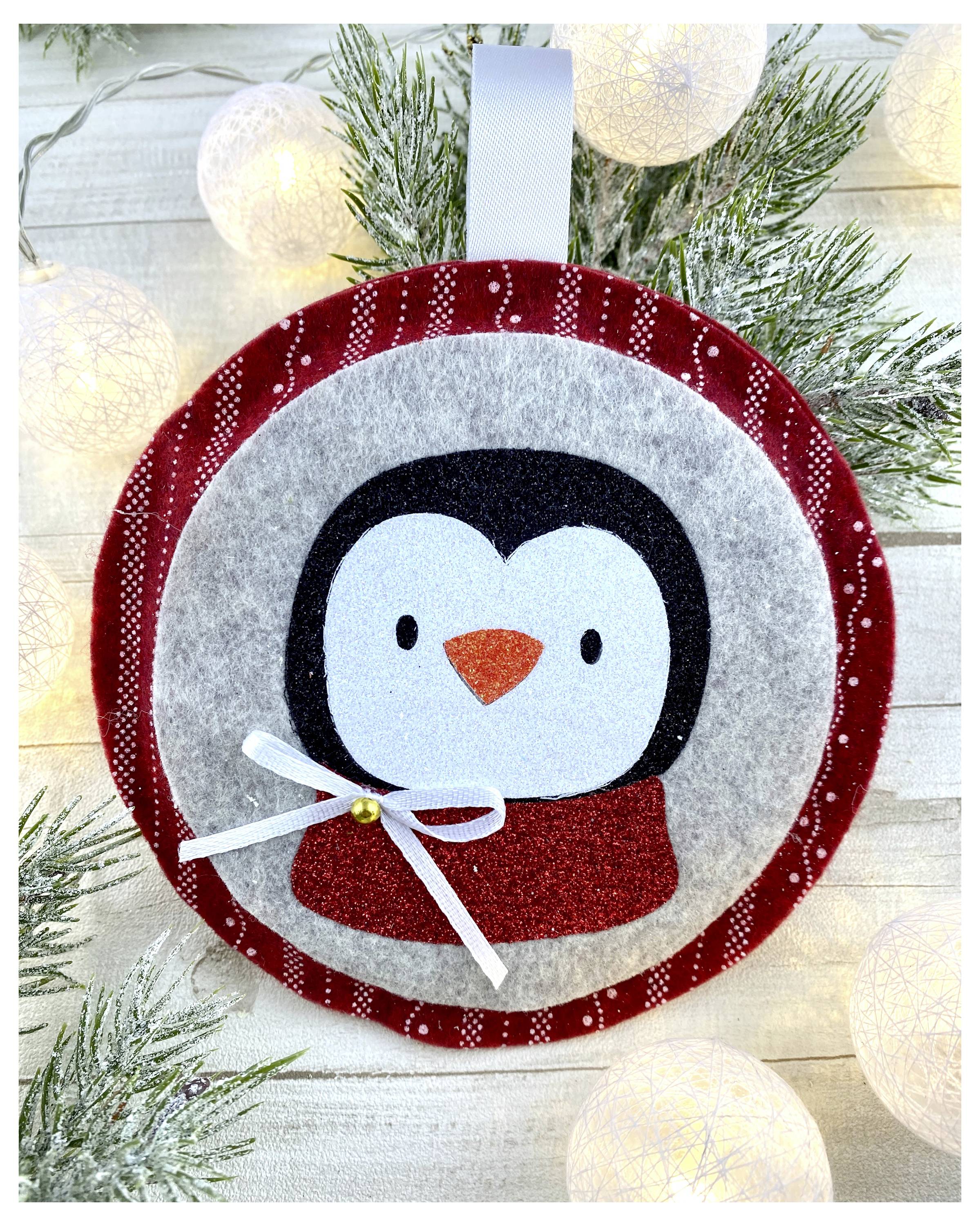 Eine Filzornament mit einem Pinguin mit rotem Schal und Schleife, umgeben von festlicher Beleuchtung und Kiefernzweigen.