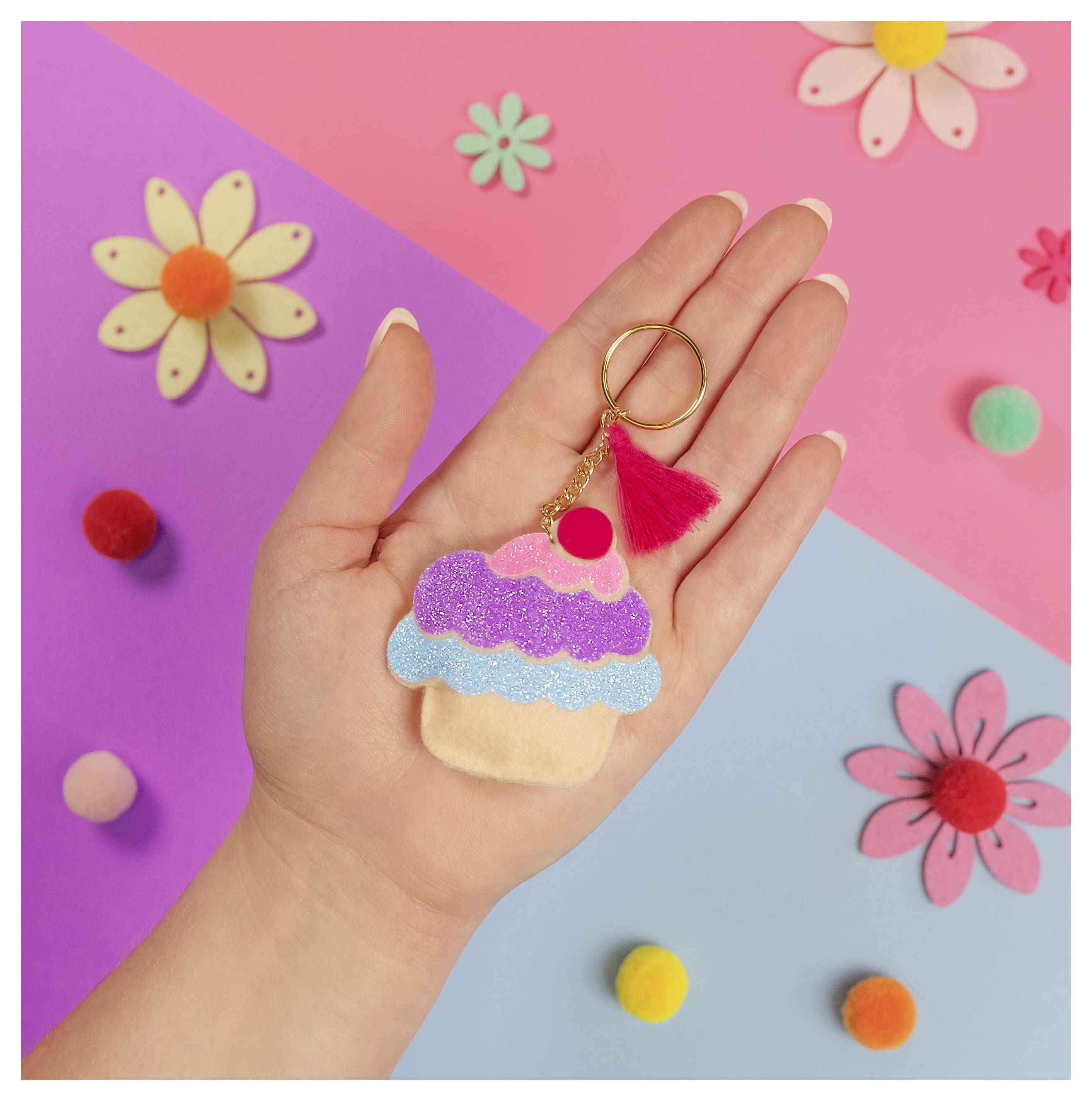 Eine Hand hält einen cupcakeförmigen Schlüsselanhänger mit rosa und blauem Glitter vor einem farbenfrohen Hintergrund mit Blumen und Pompon.