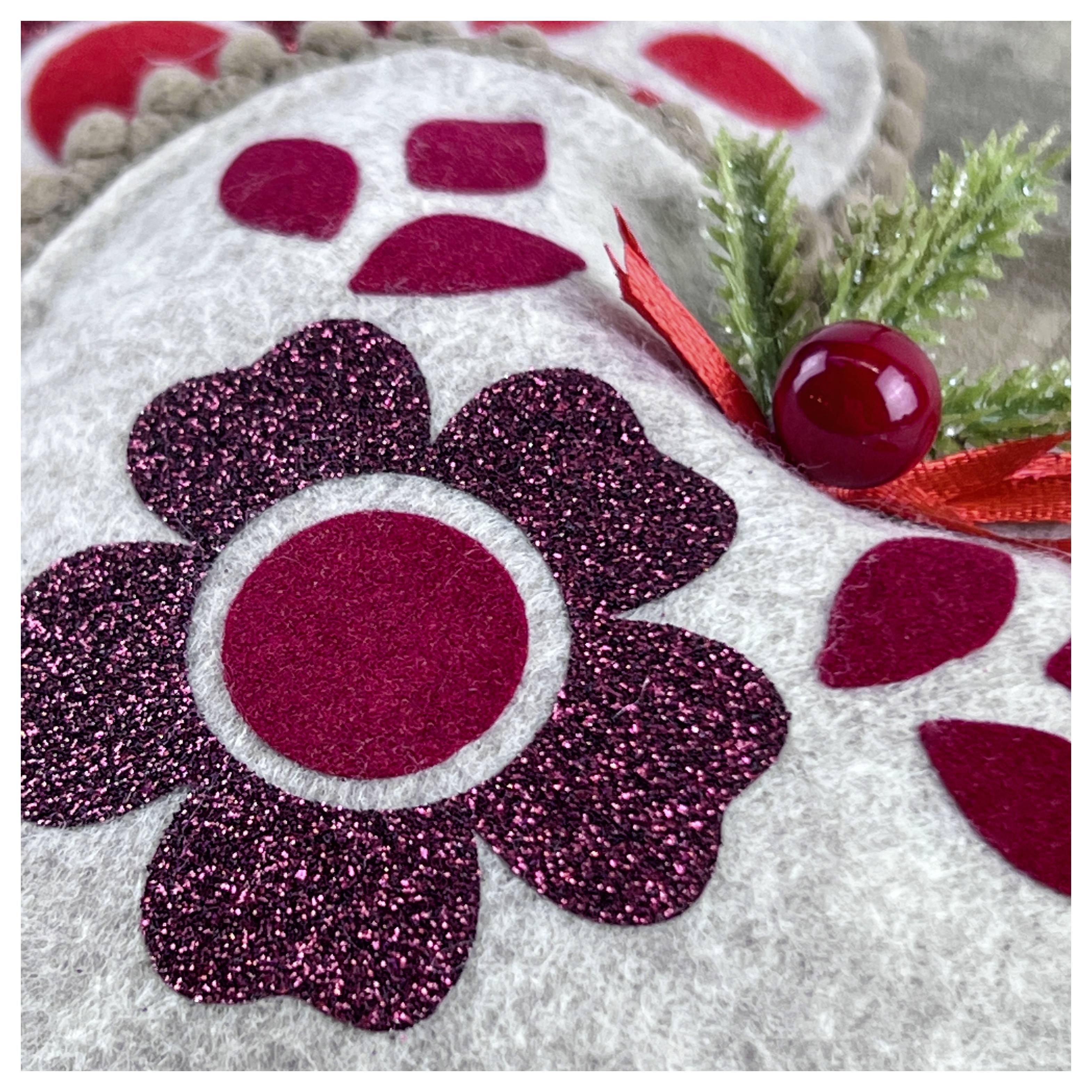 Eine Nahaufnahme eines dekorativen Weihnachtsschmucks mit einem glitzernden Blumendesign auf Filz, mit roten Akzenten und künstlichem Grün.