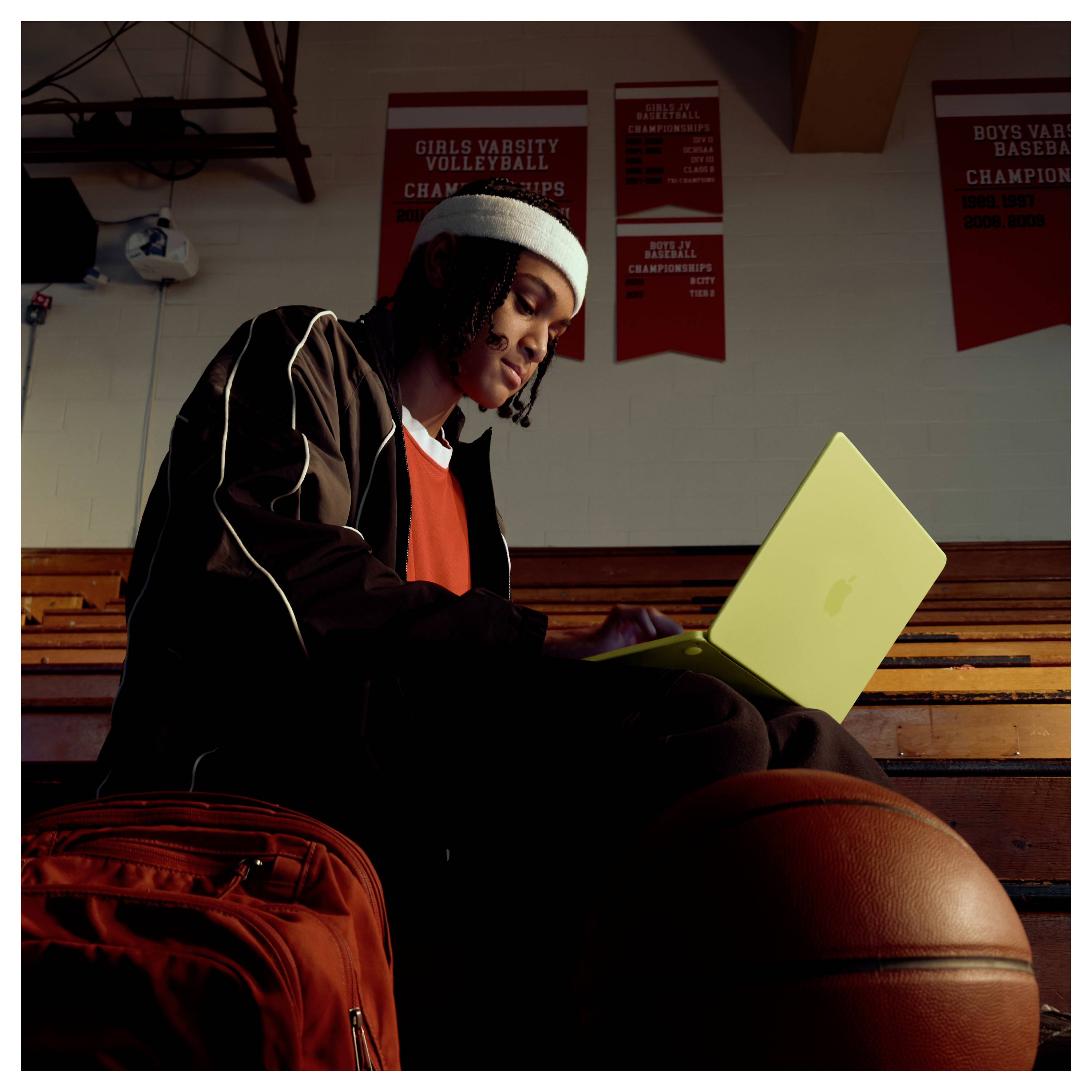 Eine Person in einer Fitnessstudio-Umgebung, sitzend auf Tribünen mit einem Laptop, Basketball und Rucksack. Meisterschaftsbanner hängen an der Wand darüber.