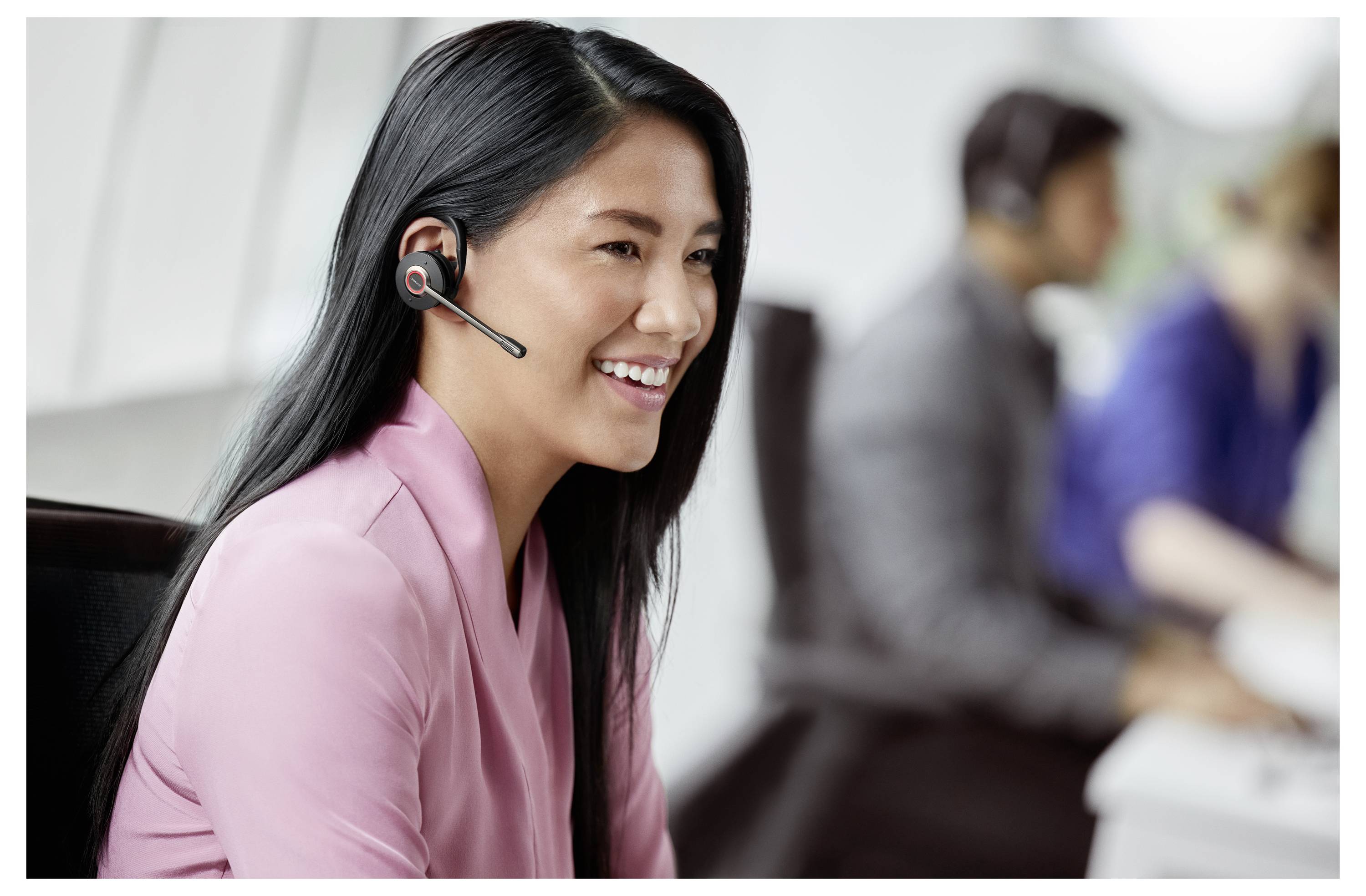 Lächelnde Kundendienstmitarbeiterin mit Headset, die an einem Schreibtisch sitzt, während Kollegen im Hintergrund verschwommen sind.