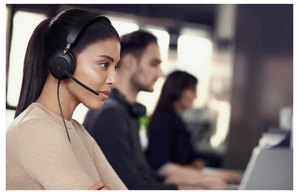 Eine Frau mit Kopfhörer arbeitet an einem Computer in einer Callcenter-Umgebung. Zwei weitere Personen sind im Hintergrund unscharf.