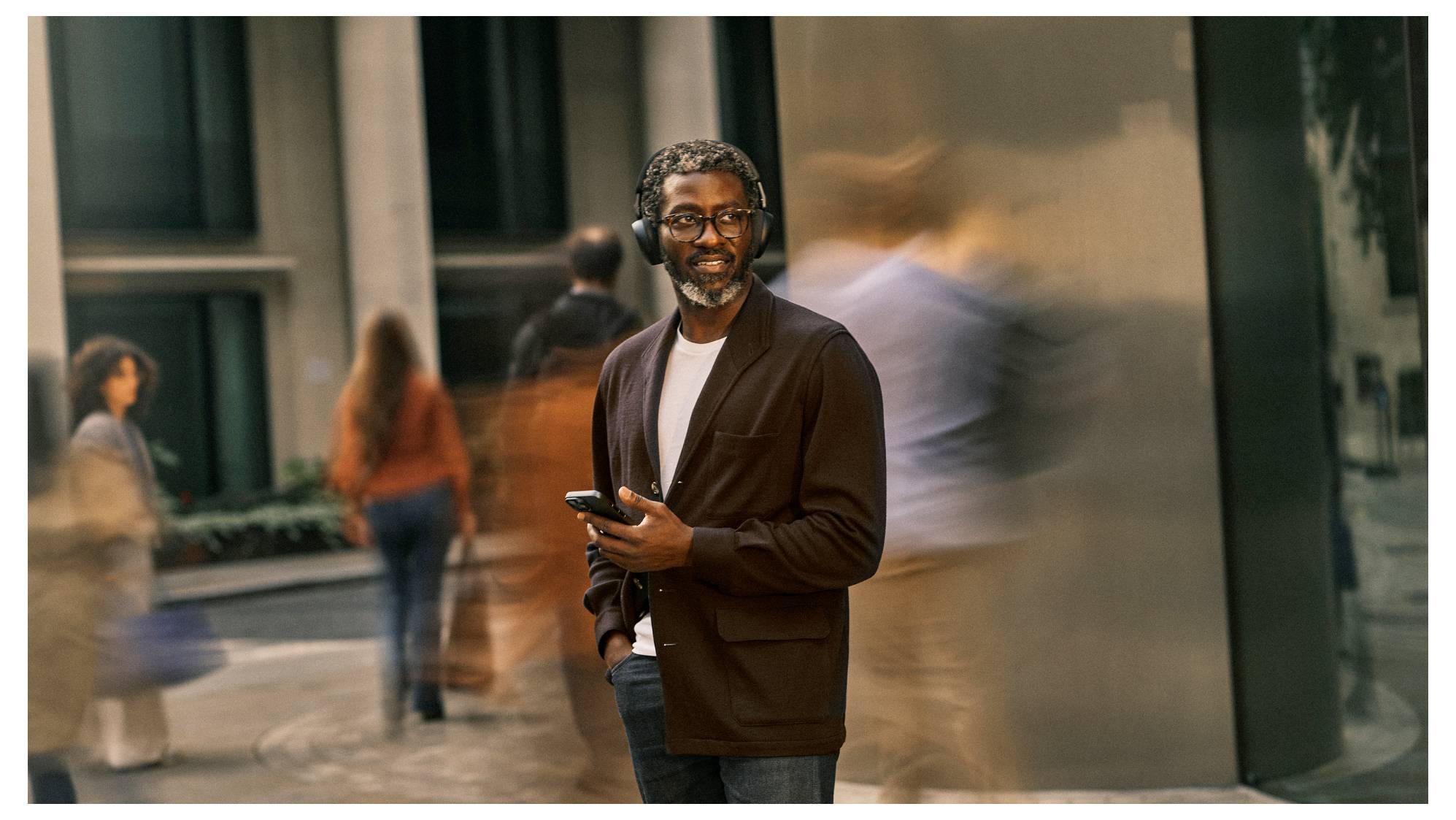 Porträt eines Mannes mit Brille, der ein Telefon hält und im Freien lächelt, mit verschwommenen Fußgängern und Stadtgebäuden im Hintergrund.