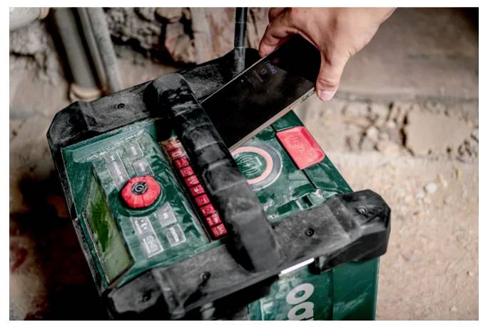 Eine staubige, grüne Autobatterie wird mit einem Multimeter oder einem ähnlichen Gerät von einer Hand einer Person getestet oder überprüft, was auf Wartung hindeutet.