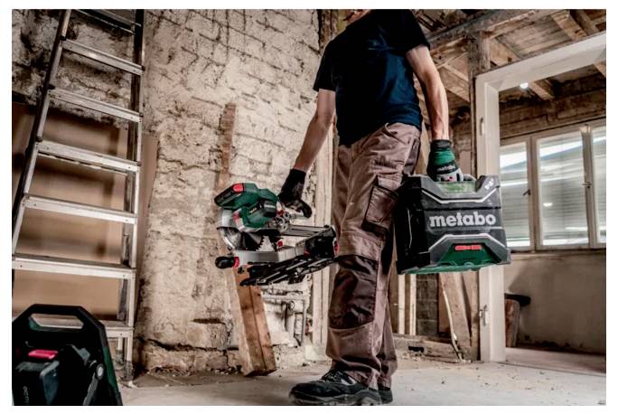 Eine Person in Schutzausrüstung trägt einen 'Metabo' Elektrowerkzeugkoffer und ein Werkzeug in einem unfertigen Raum mit einer Leiter und freigelegten Ziegelwänden.