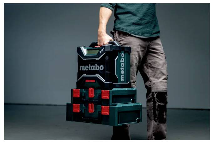 Eine Person, die einen Stapel von Metabo-Werkzeugkästen mit einem Griff trägt, trägt Arbeitshosen und zeigt Portabilität und einfachen Transport.
