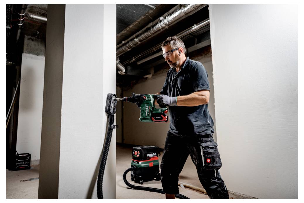 Eine Holzplanke ist auf einer perforierten Werkbank mit zwei Spannzwingen befestigt. Eine Spannzwinge ist grün und aufrecht; die andere ist silbern und flach angebracht.