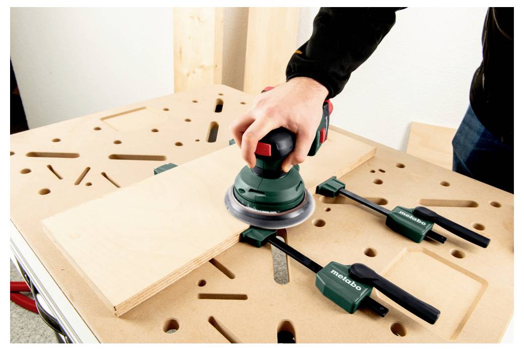 Eine Person benutzt einen Elektroschleiffer auf einem Holzbrett, das mit Zwingen auf einer Werkbank gesichert ist.