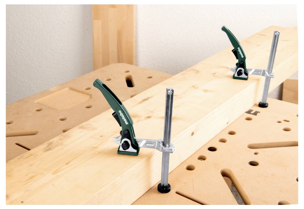 Ein Holzbrett, das mit zwei Metallzwingen auf einer Werkbank befestigt ist, zeigt eine Anordnung zum Holzbearbeiten oder Handwerk.