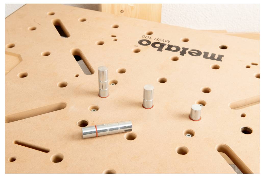 Eine Arbeitsplatte aus Holz mit verschiedenen Löchern und vier eingefügten zylindrischen Metallstiften. Das Logo „metabo