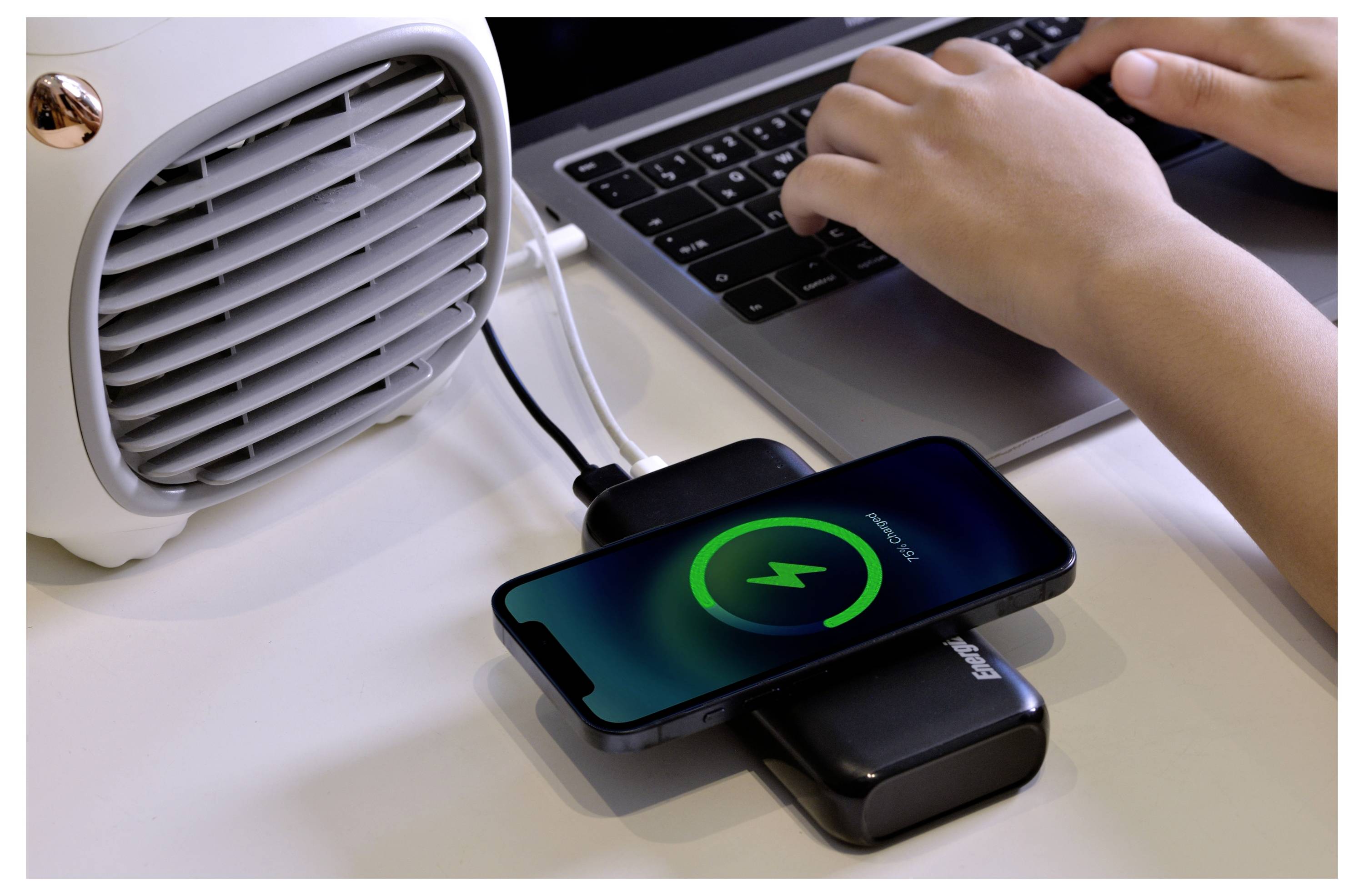 Eine Person tippt auf einem Laptop, während ein Smartphone neben einer kleinen Heizung auf einem Schreibtisch drahtlos aufgeladen wird.