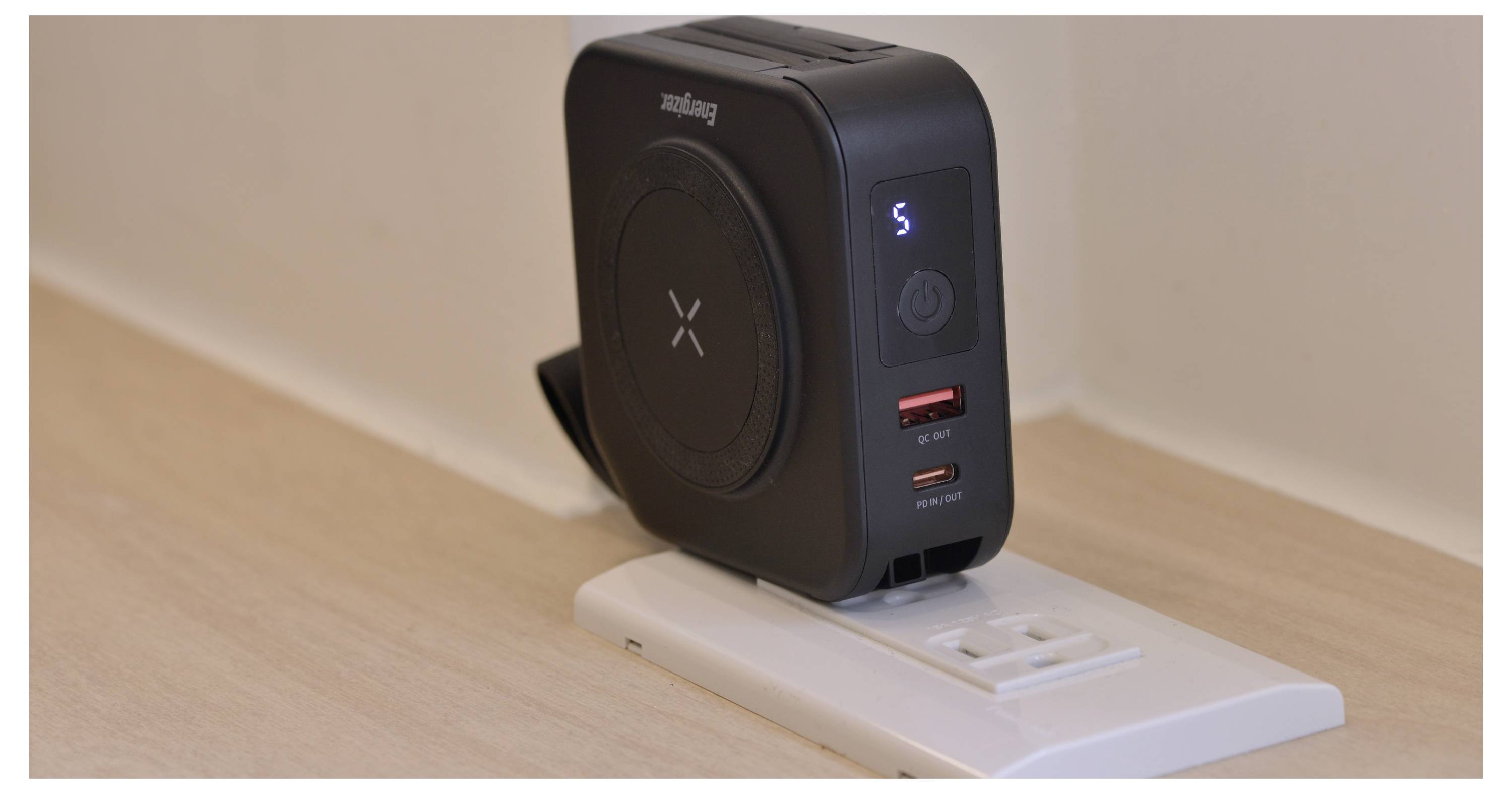 Ein schwarzes kabelloses Ladegerät und ein Netzteil, das in eine Wandsteckdose eingesteckt ist, mit LED-Anzeigen und zwei USB-Anschlüssen auf einer hellen Holzoberfläche.