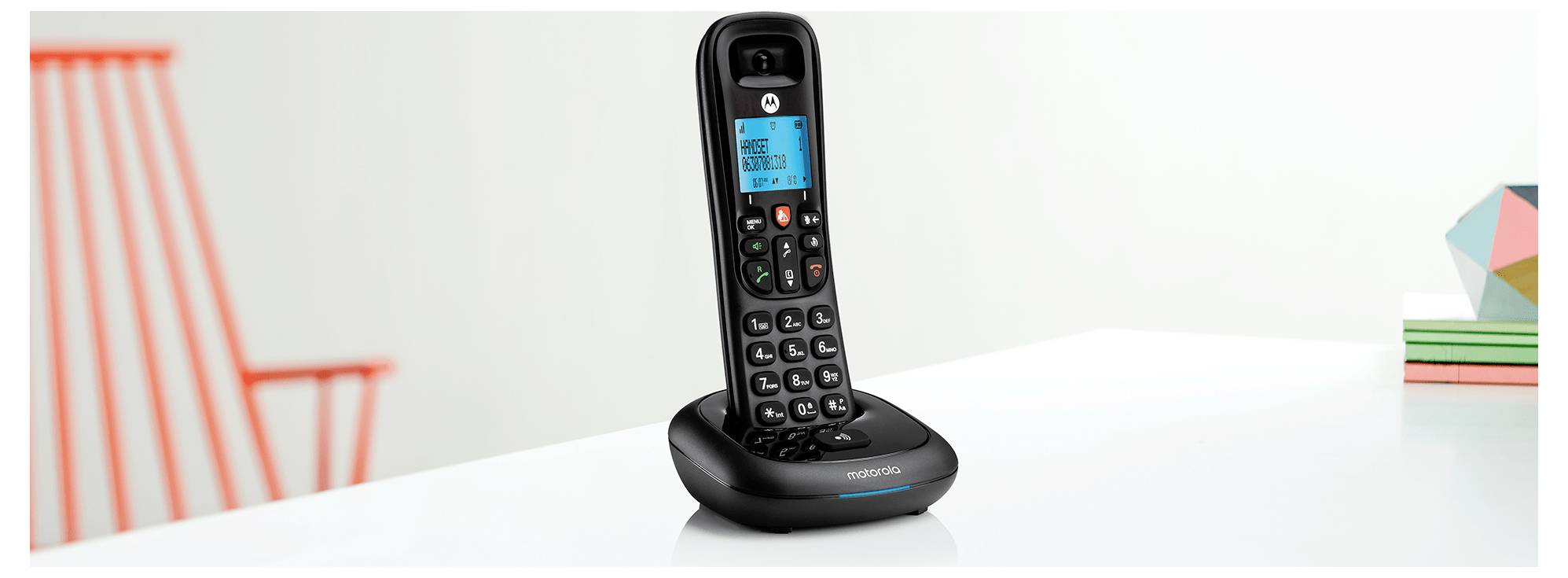 Ein schwarzes schnurloses Telefon mit digitalem Display auf seiner Basis, auf einem weißen Tisch neben bunten geometrischen Dekorationsobjekten platziert.