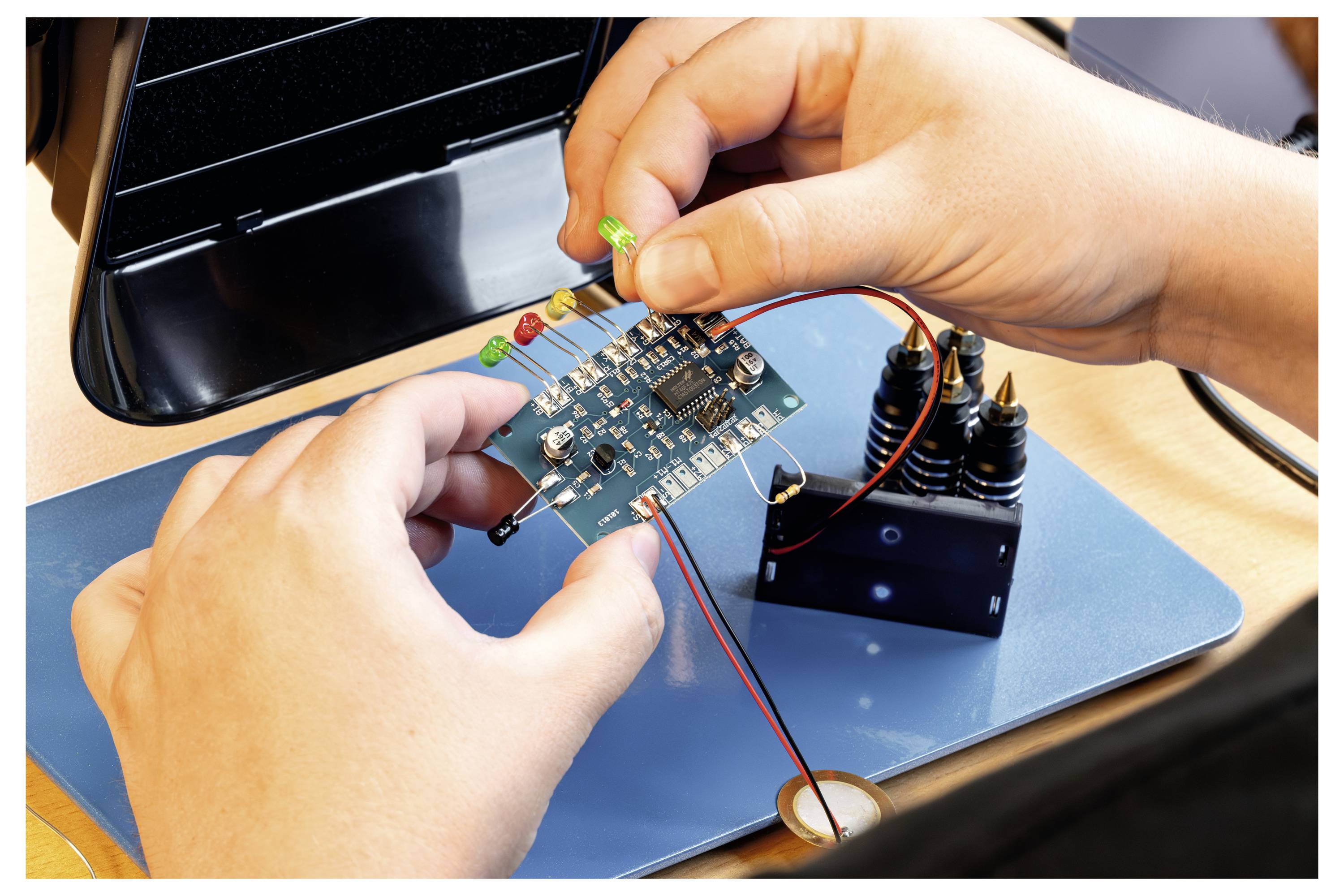 Zwei Hände montieren eine elektronische Leiterplatte mit Komponenten, einschließlich LEDs, auf einer Tischarbeitsfläche.